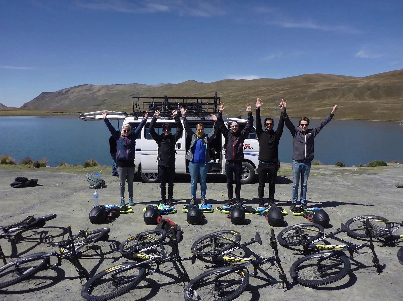 Travelers raising arms near mountain bikes and a support van by a lake.