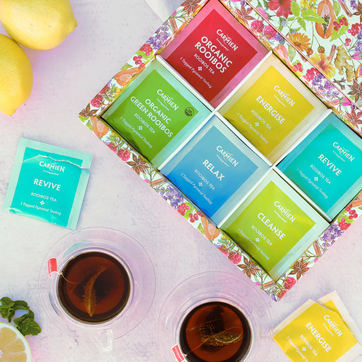 A gift box open to reveal Carmién tea bags. The box is standing on a white counter surrounded by cups of tea and cut and uncut lemons.
