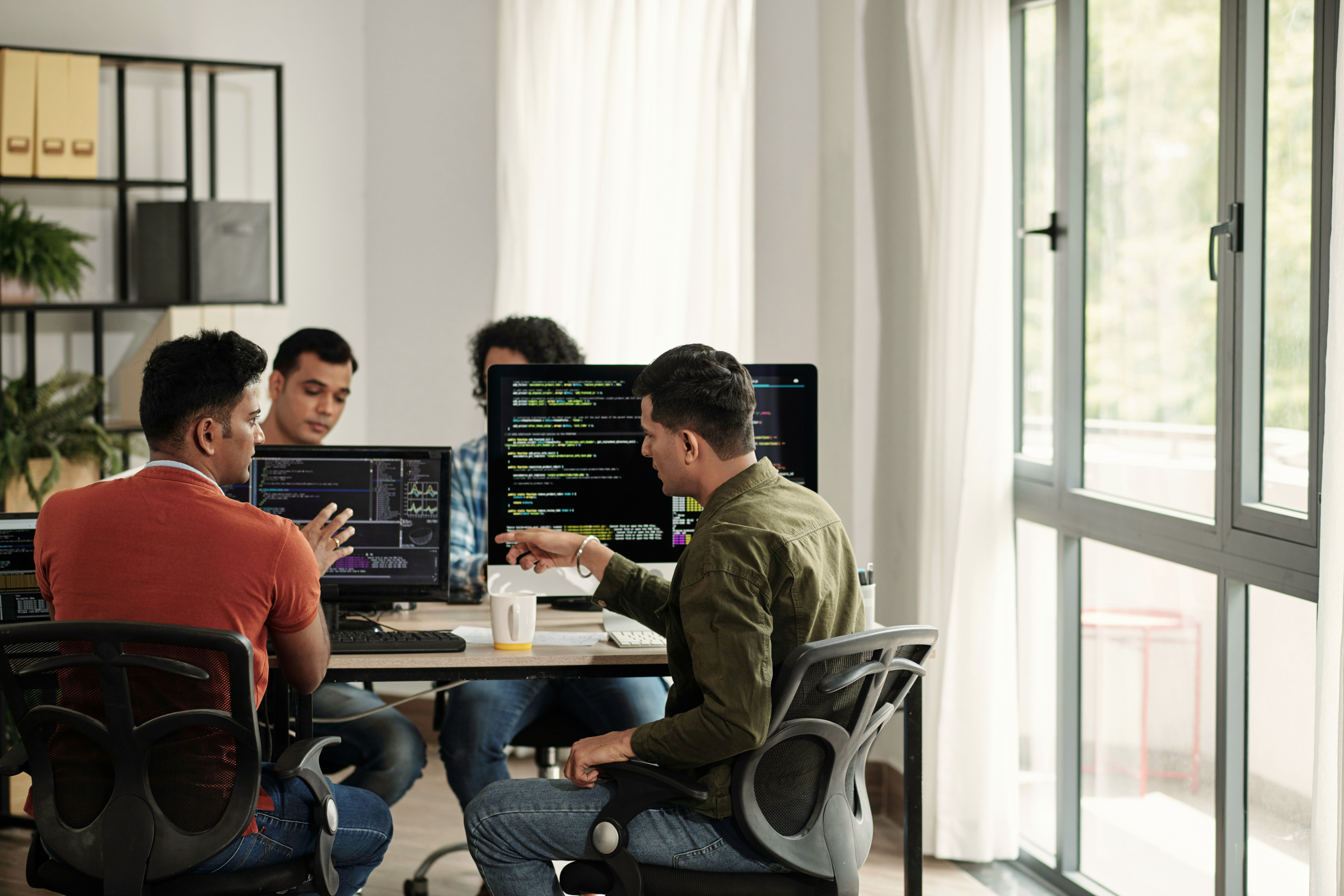 Three individuals collaborate around a table with laptops in a bright, modern workspace.