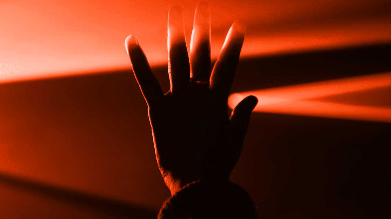 Silhouette of a raised hand, reaching toward bright orange light beams in a dark, atmospheric scene