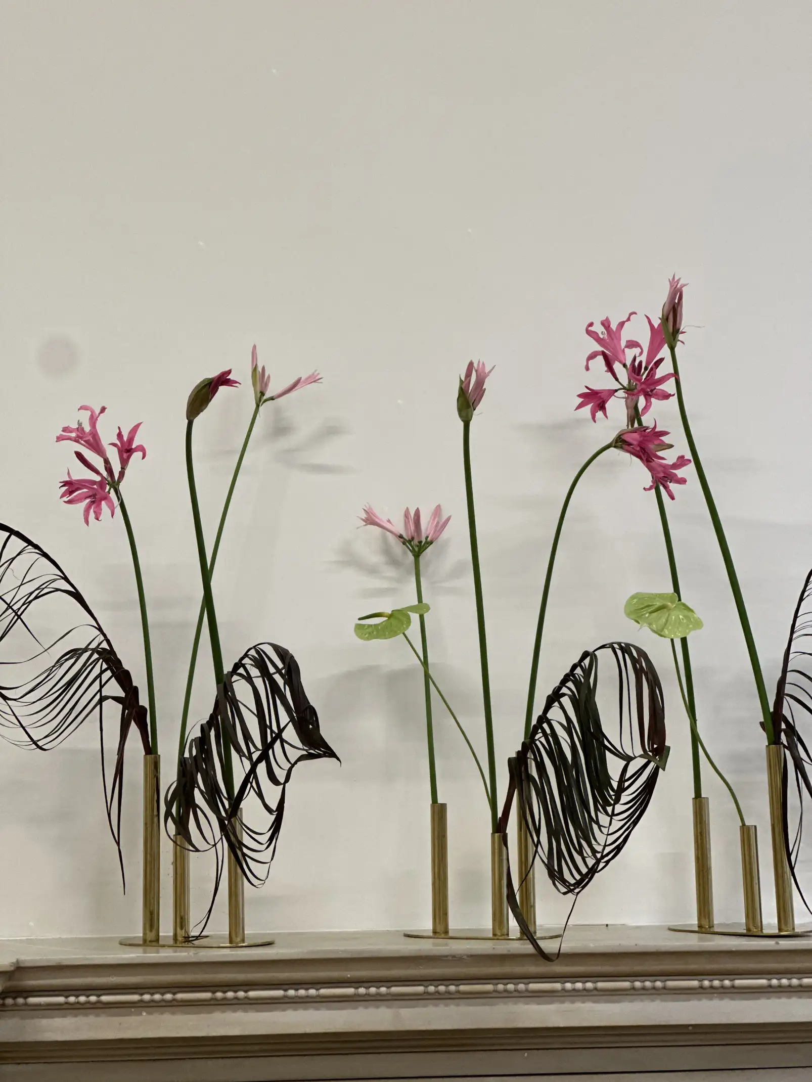 Arreglo minimalista con flores rosas de tallo largo y hojas oscuras recortadas, colocadas en jarrones cilíndricos dorados alineados sobre una repisa blanca.