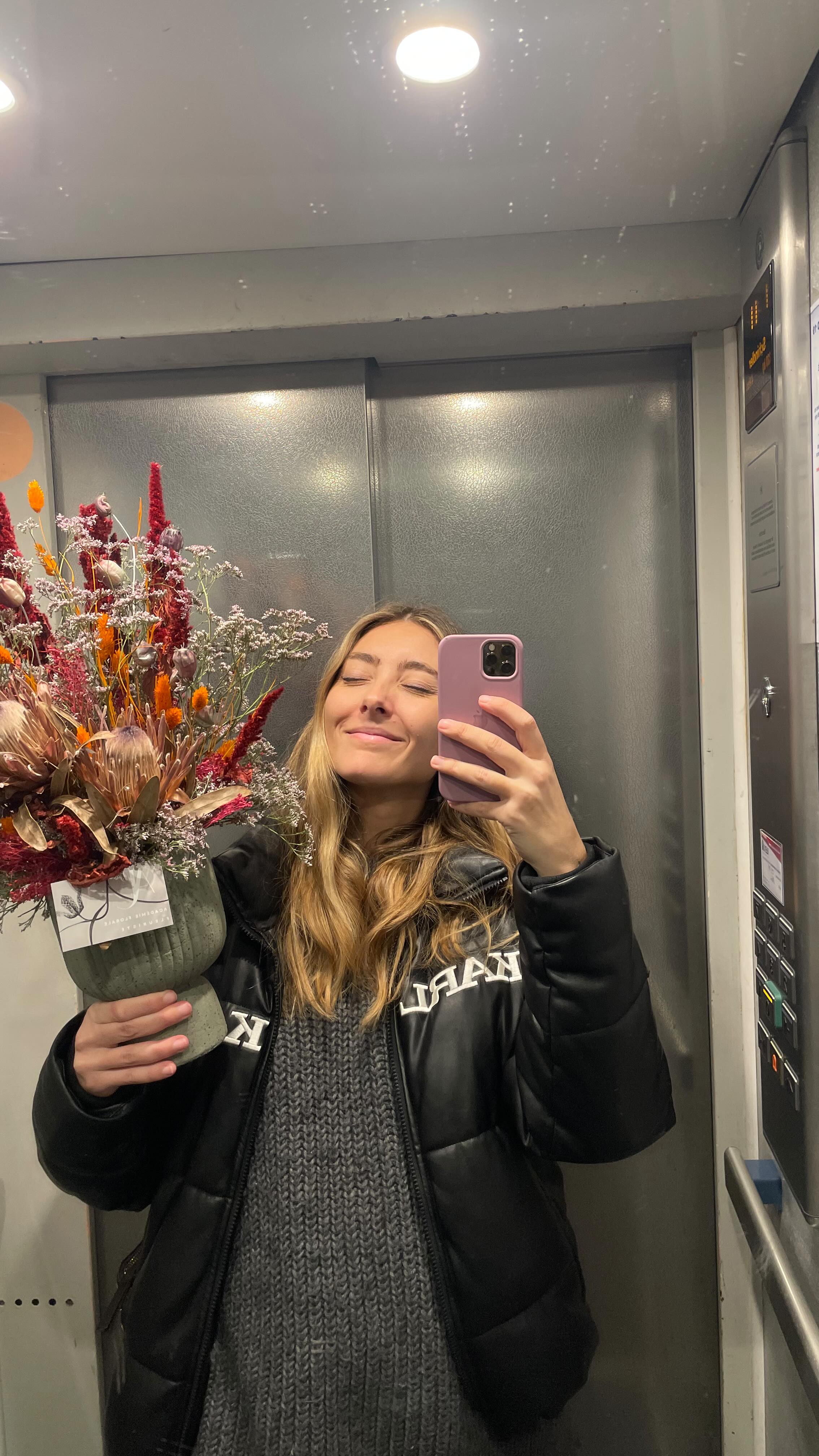 Young person with wavy hair poses confidently in sunlight against a modern building backdrop. Wears a black blazer and silver necklace, conveying elegance.