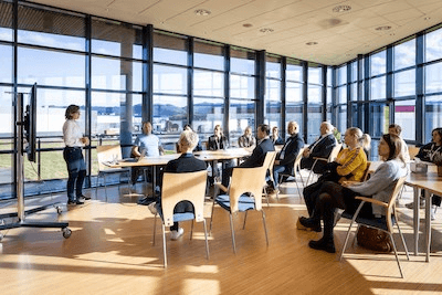 A group of people seated in a bright conference room, listening to a presenter near a whiteboard.