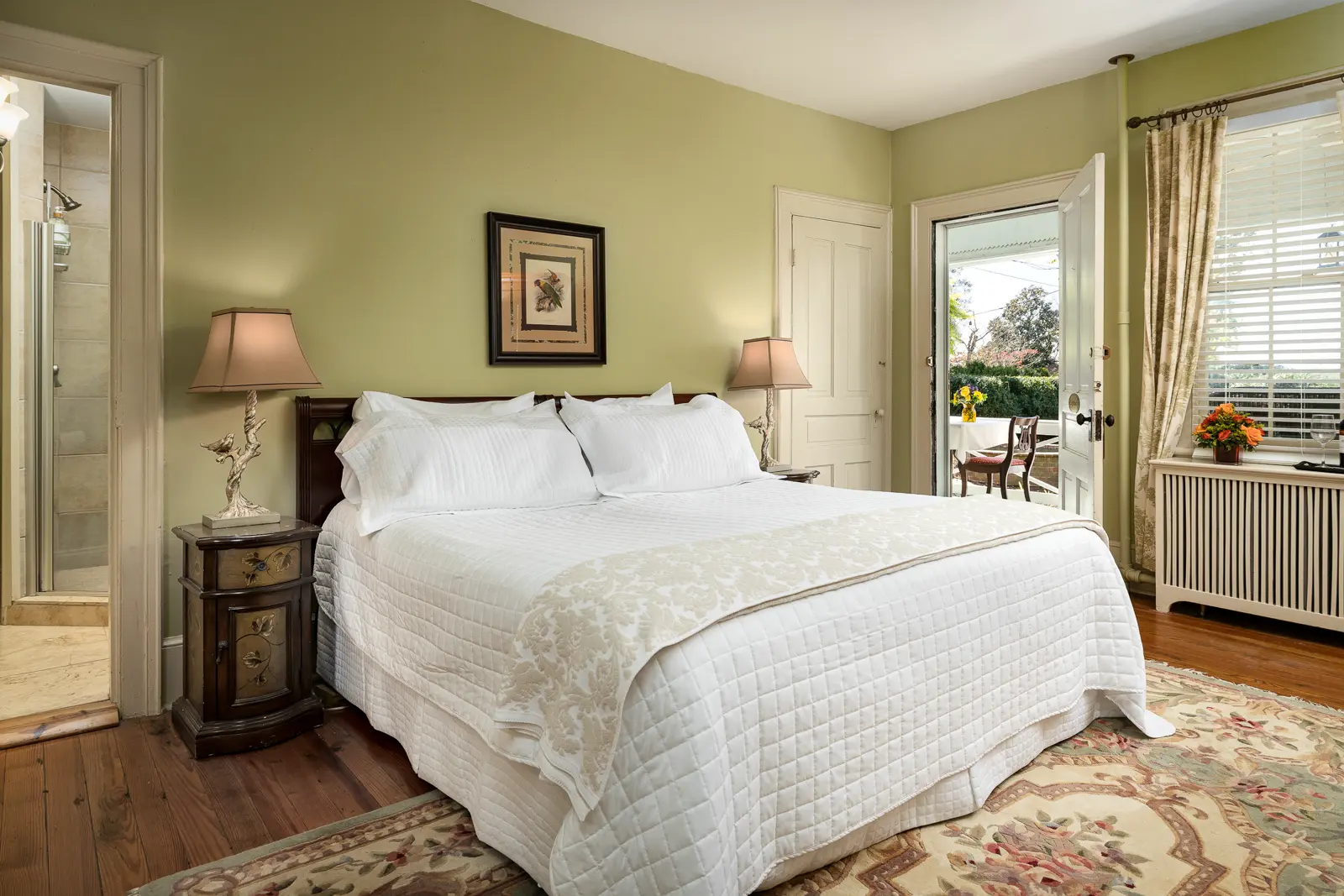 A charming, rustic white-brick cottage at The Estate at Meander features a black metal roof, dark-framed windows, and a black door, accented by vibrant red flowers and warm, ambient lighting.