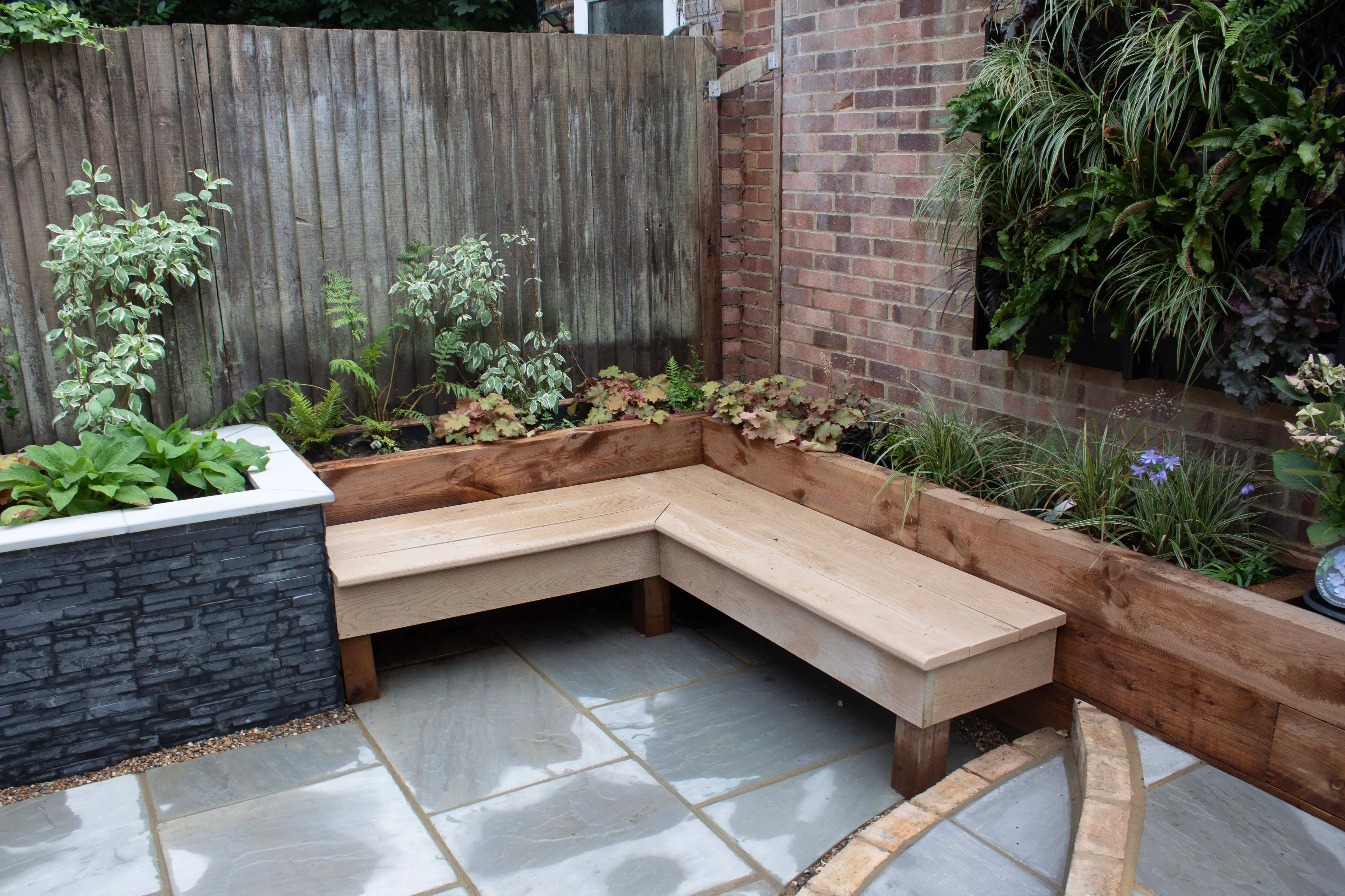 A cozy outdoor seating area with a wooden corner bench and greenery in a stylish, urban garden setting.