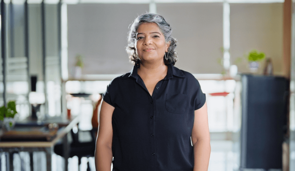 A middle-aged woman with grey-streaked hair and a black short-sleeved shirt stands confidently in a bright, modern office environment, smiling gently at the camera.