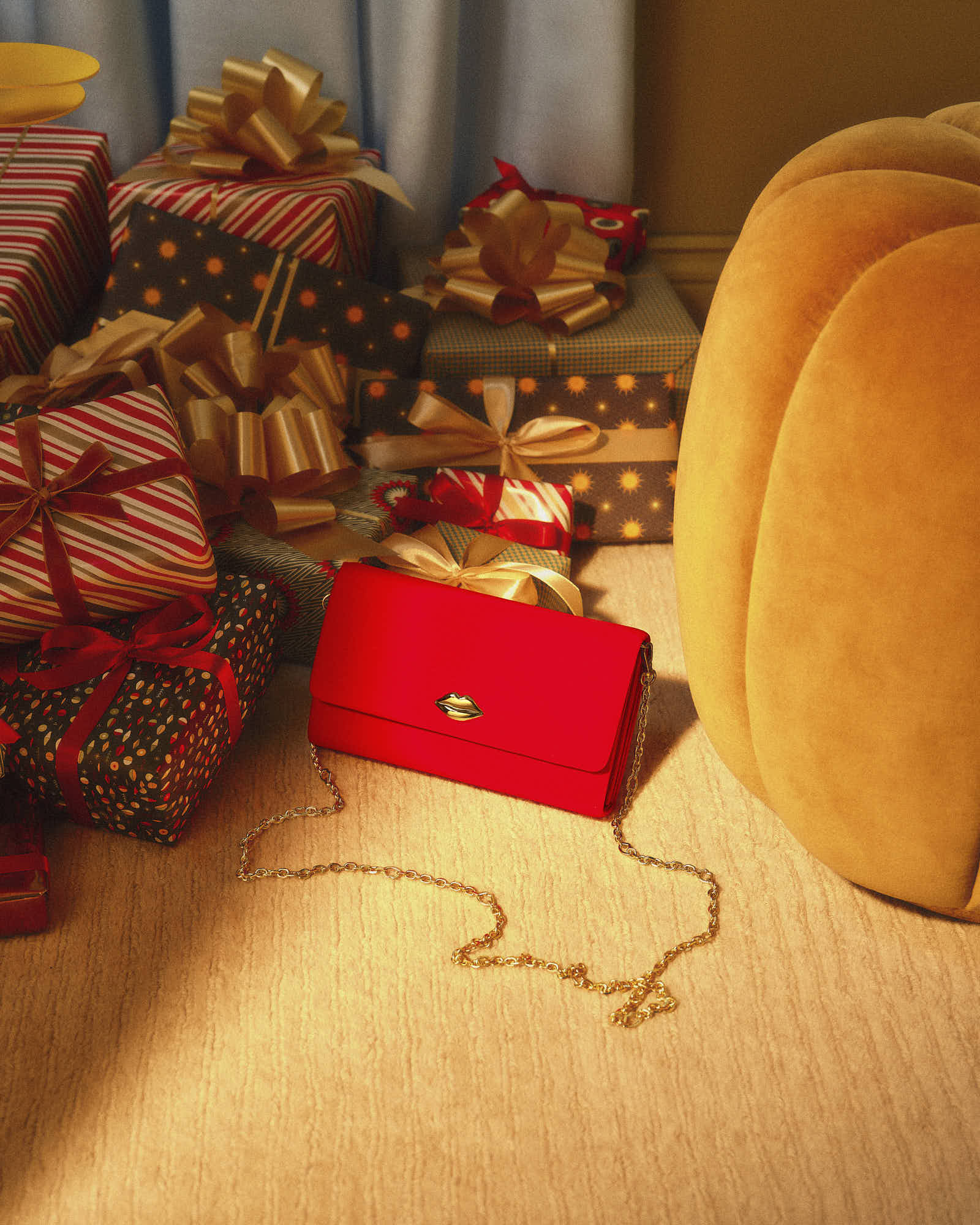 Red handbag with gold chain among wrapped gifts and plush orange chair.