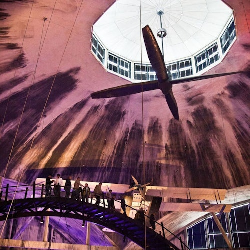 Emberek sétálnak egy emelt hídon egy nagy kupolás mennyezet alatt, középen egy világítótesttel és látható szerkezeti gerendákkal.