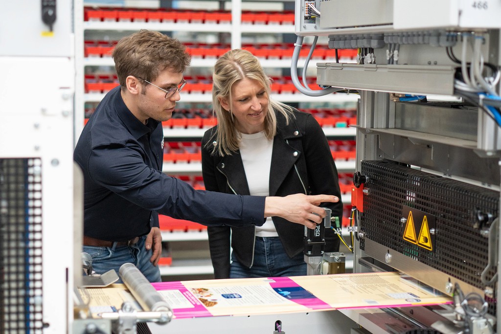 Zwei Personen inspizieren industrielle Druckmaschine. Fokus auf Qualitätskontrolle und Herstellung.