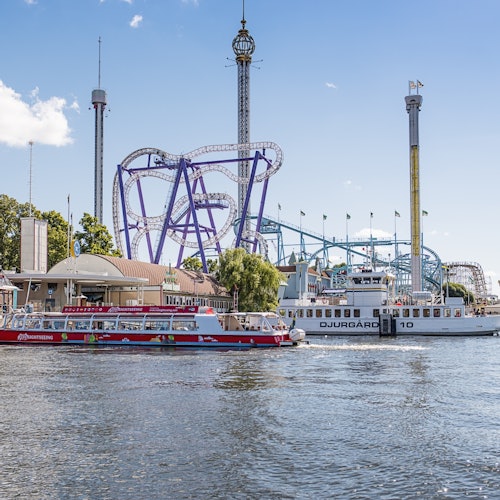 Amusement park scene with roller coasters and tall rides in the background, and boats on the water in the foreground under a blue sky.
