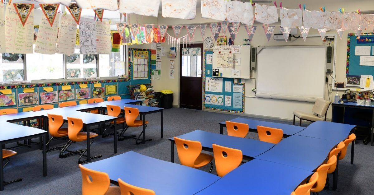 A bright primary school classroom with arranged desks, orange chairs, educational posters on the walls and children’s artwork hanging overhead.