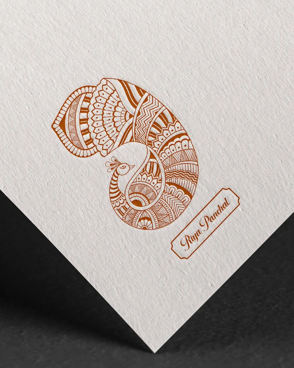 Intricate brown henna-style peacock and paisley design on a textured white card, with a small decorative greeting label, against a dark background.