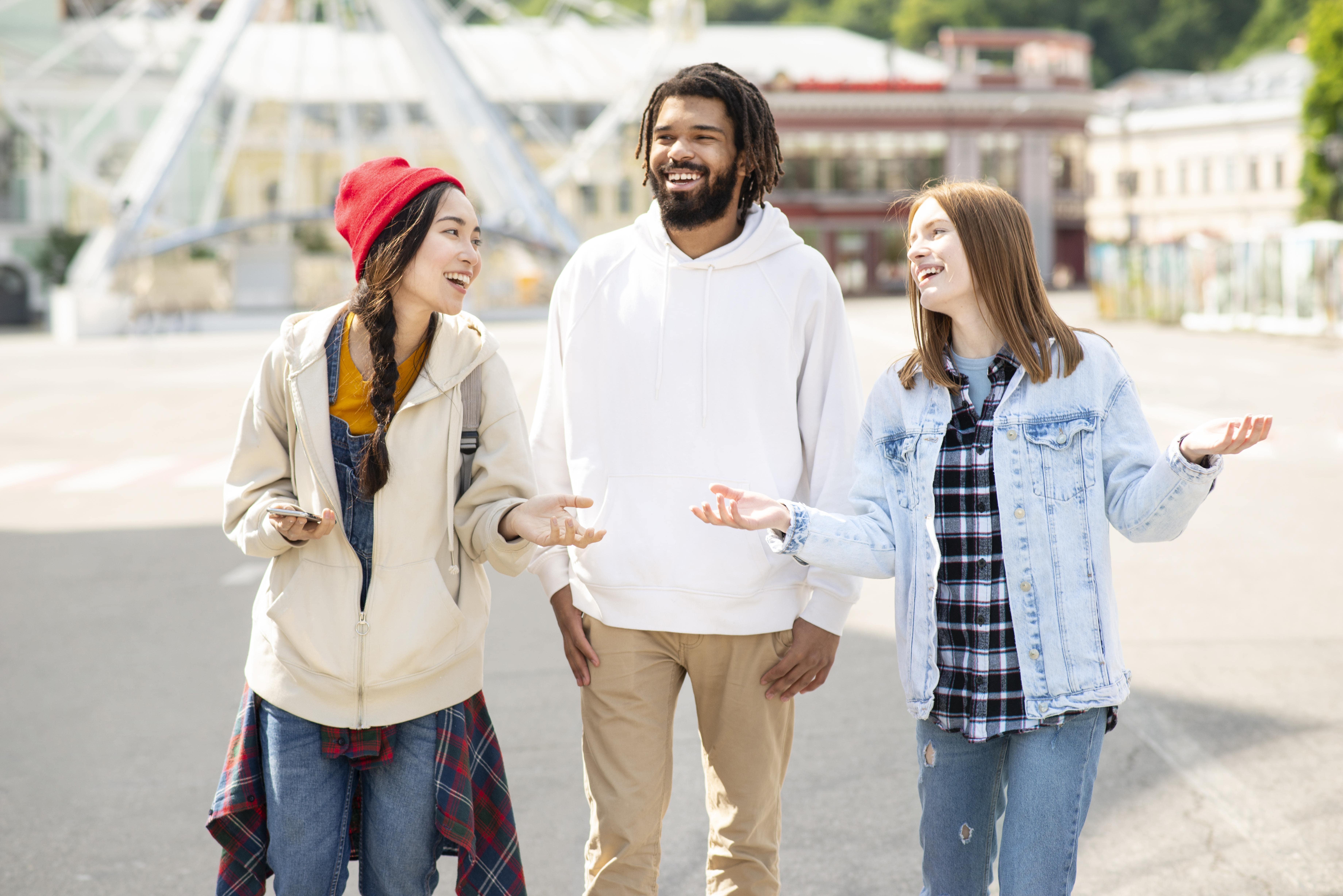 Three friends outdoors, smiling and chatting, dressed casually in a sunny setting.