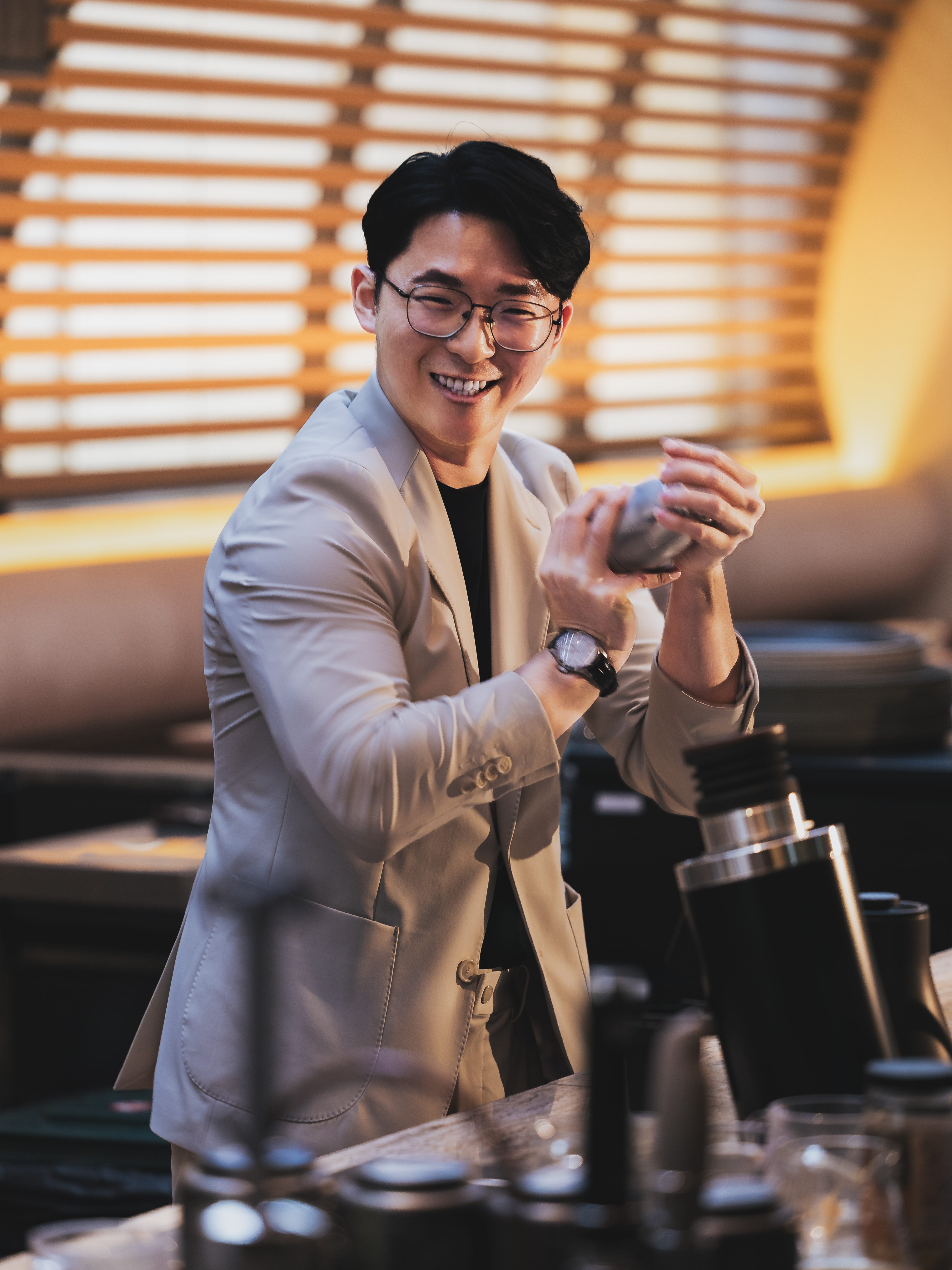 A bartender in a light grey suit joyfully shaking a cocktail shaker at a bar, with warm ambient lighting in the background.
