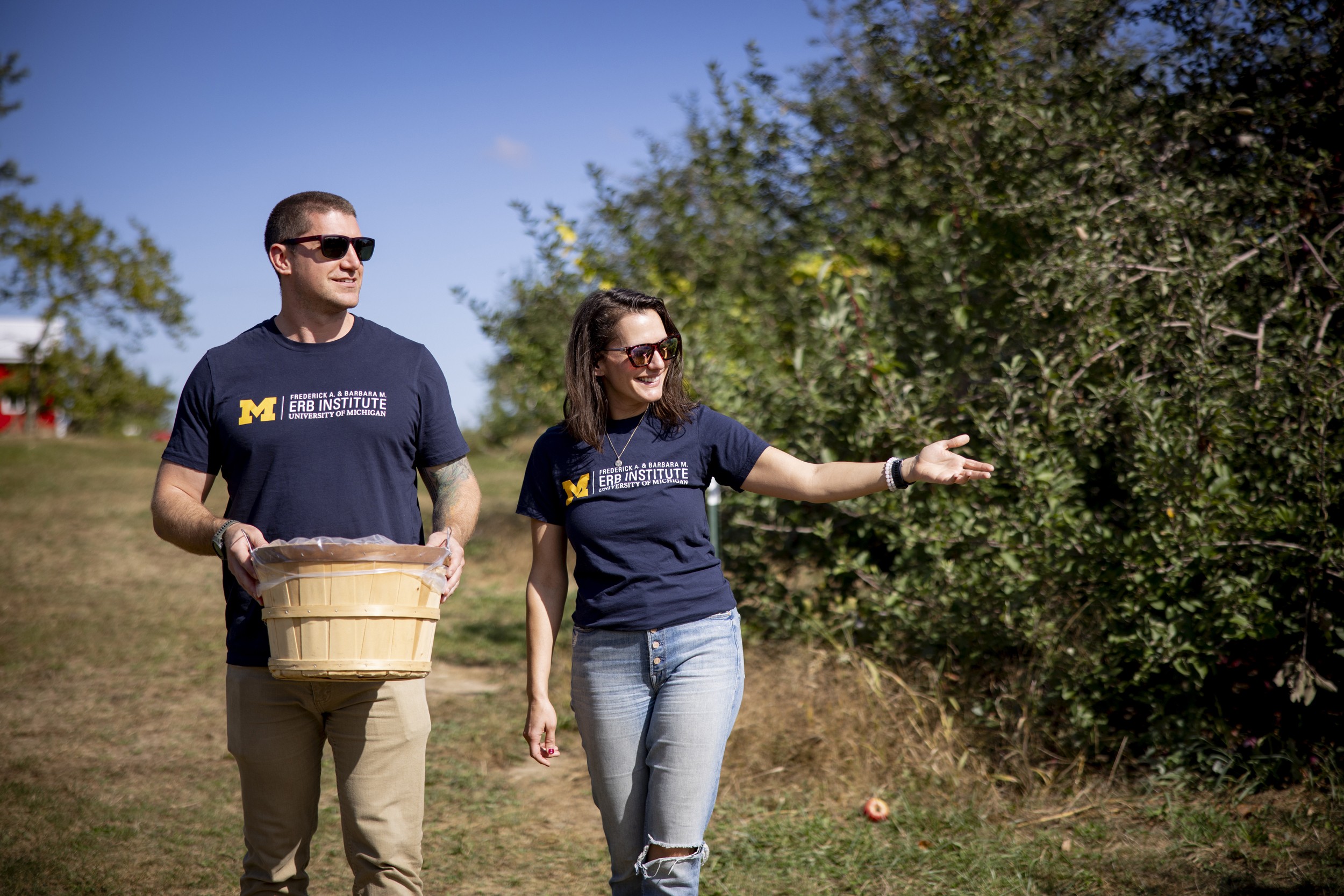 Two people wearing University of Michigan shirts walking in a sunny orchard.