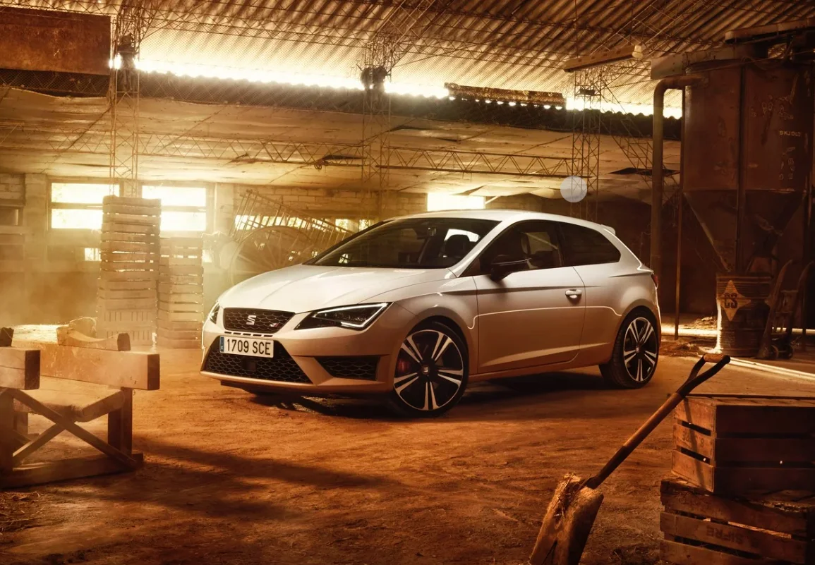 Une SEAT Leon mise en scène dans un grand hangar baigné d’une lumière chaude. La poussière au sol et les éléments en bois soulignent le contraste entre l’environnement brut et le design moderne de la compacte sportive.