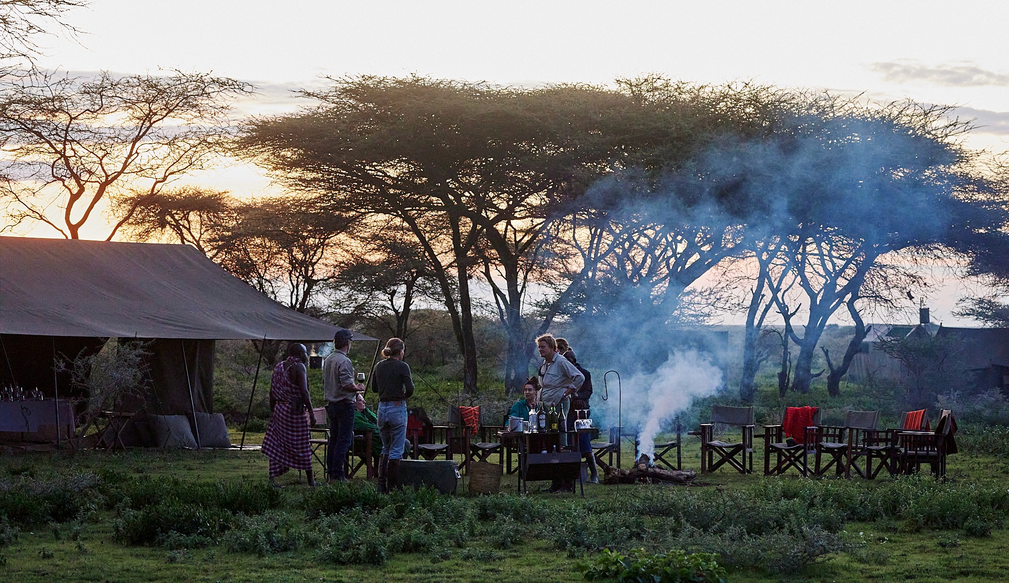 Kilimanjaro Elephant Ride, Arusha, Tanzania. Människor står runt en lägereld djup i Tanzanias vildmark. Tält står uppsatta och det är skymning.
