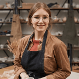A woman wearing an apron stands with a bowl of clay, ready to start her pottery work in a well-lit studio.