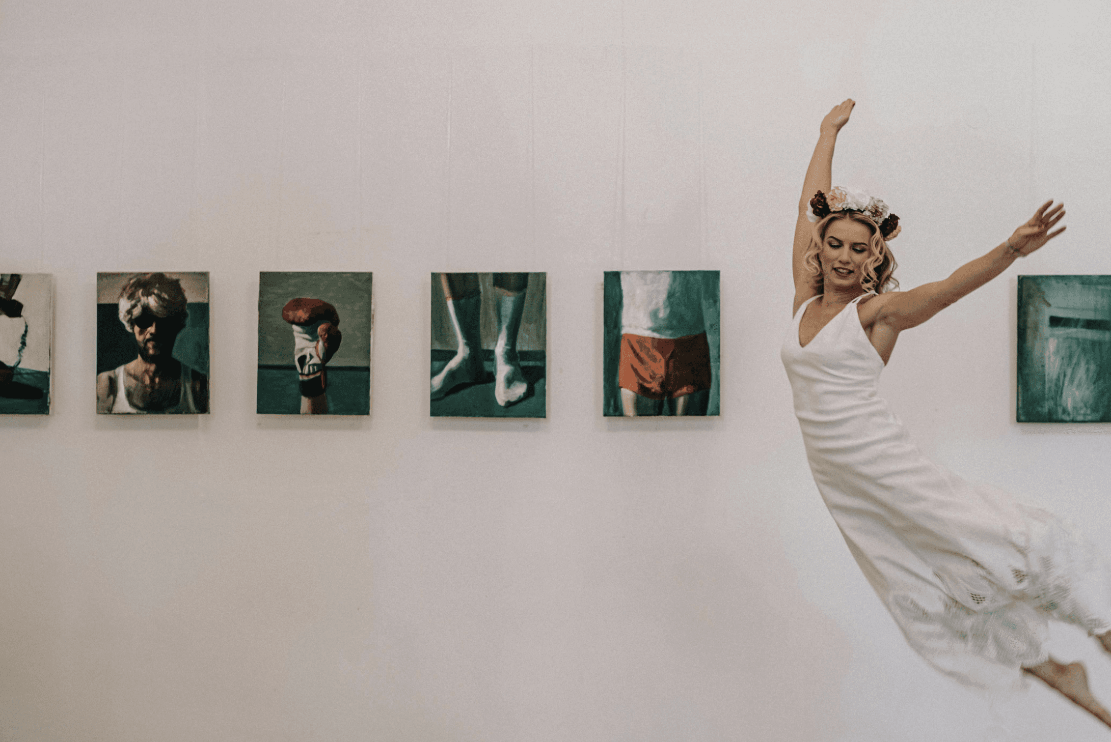Woman in a white dress and flower crown leaping in an art gallery, with square paintings on the wall.