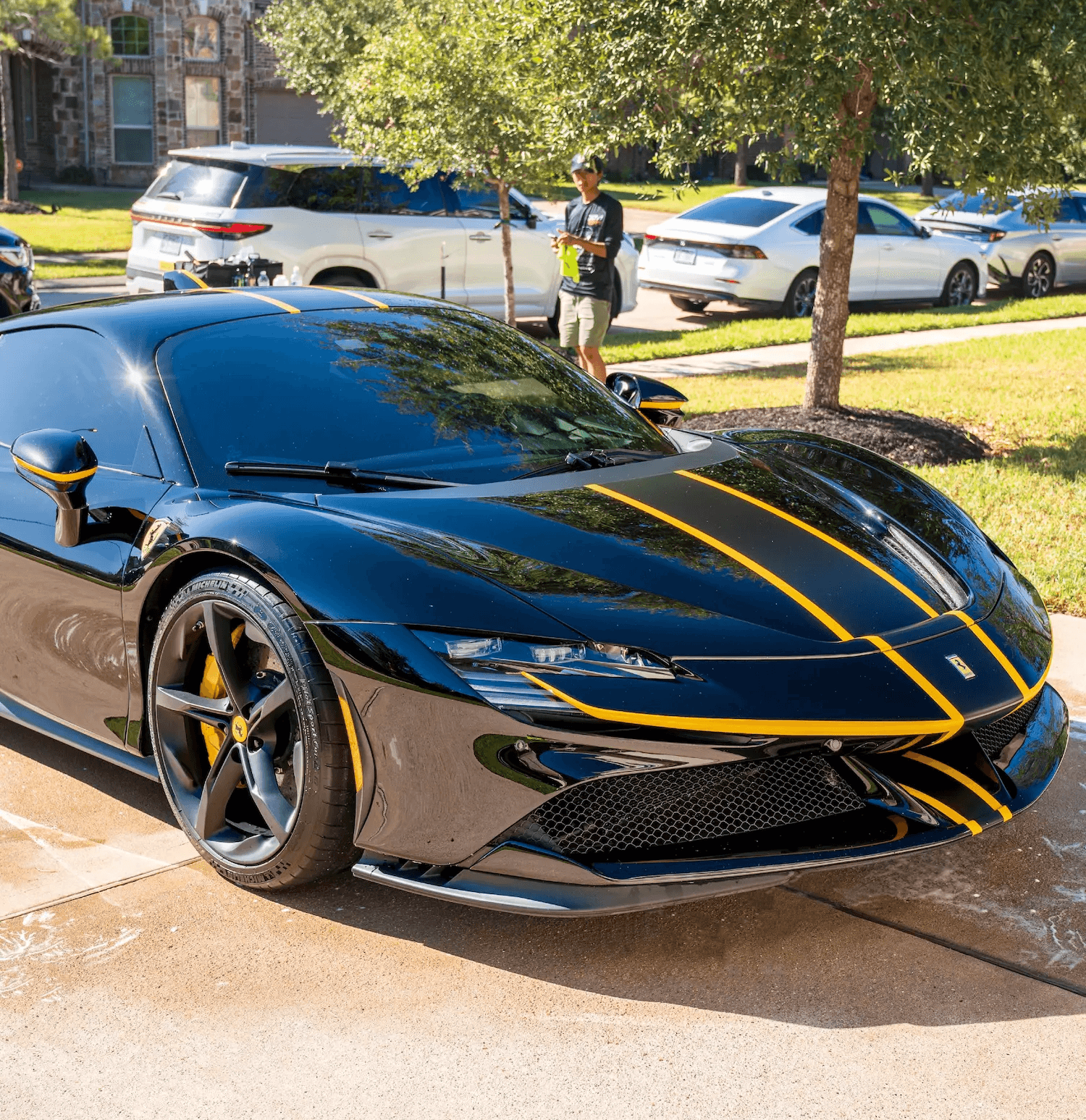 A highly reflective black Ferrari with yellow racing stripes parked on a wet residential driveway after a detailing service.