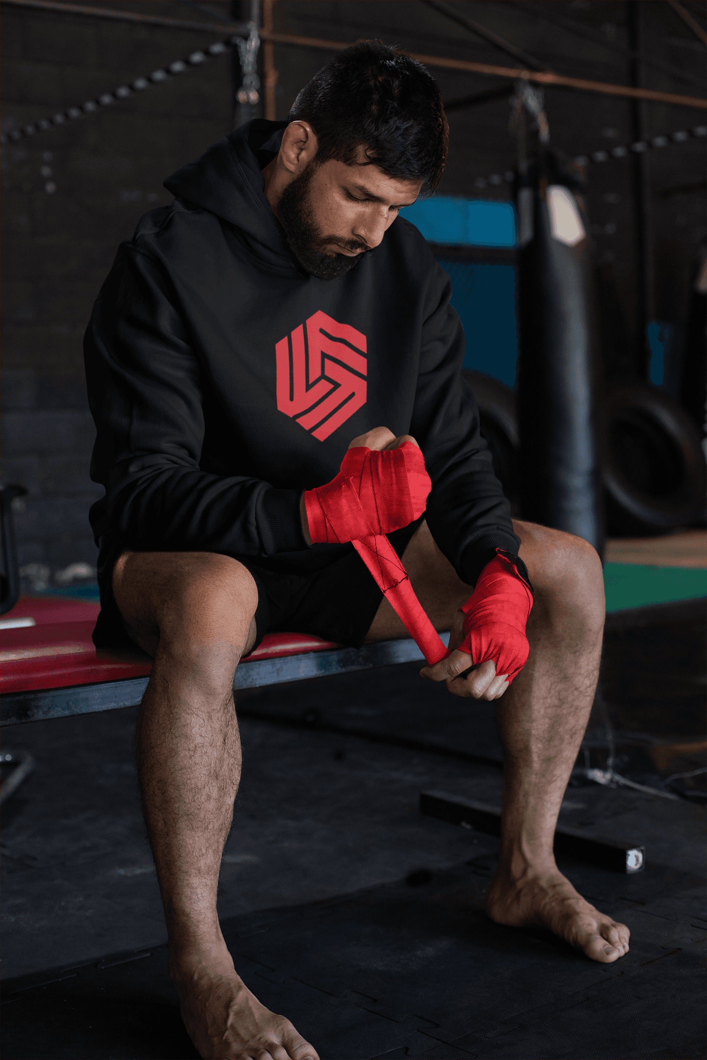 A focused man sitting on a bench in a training gym, wrapping his hands with red hand wraps. He is wearing a black hoodie that features a bold red hexagonal logo representing 'Flexibility For Fighters.' The background shows gym equipment such as punching bags and ropes, creating an authentic training environment.