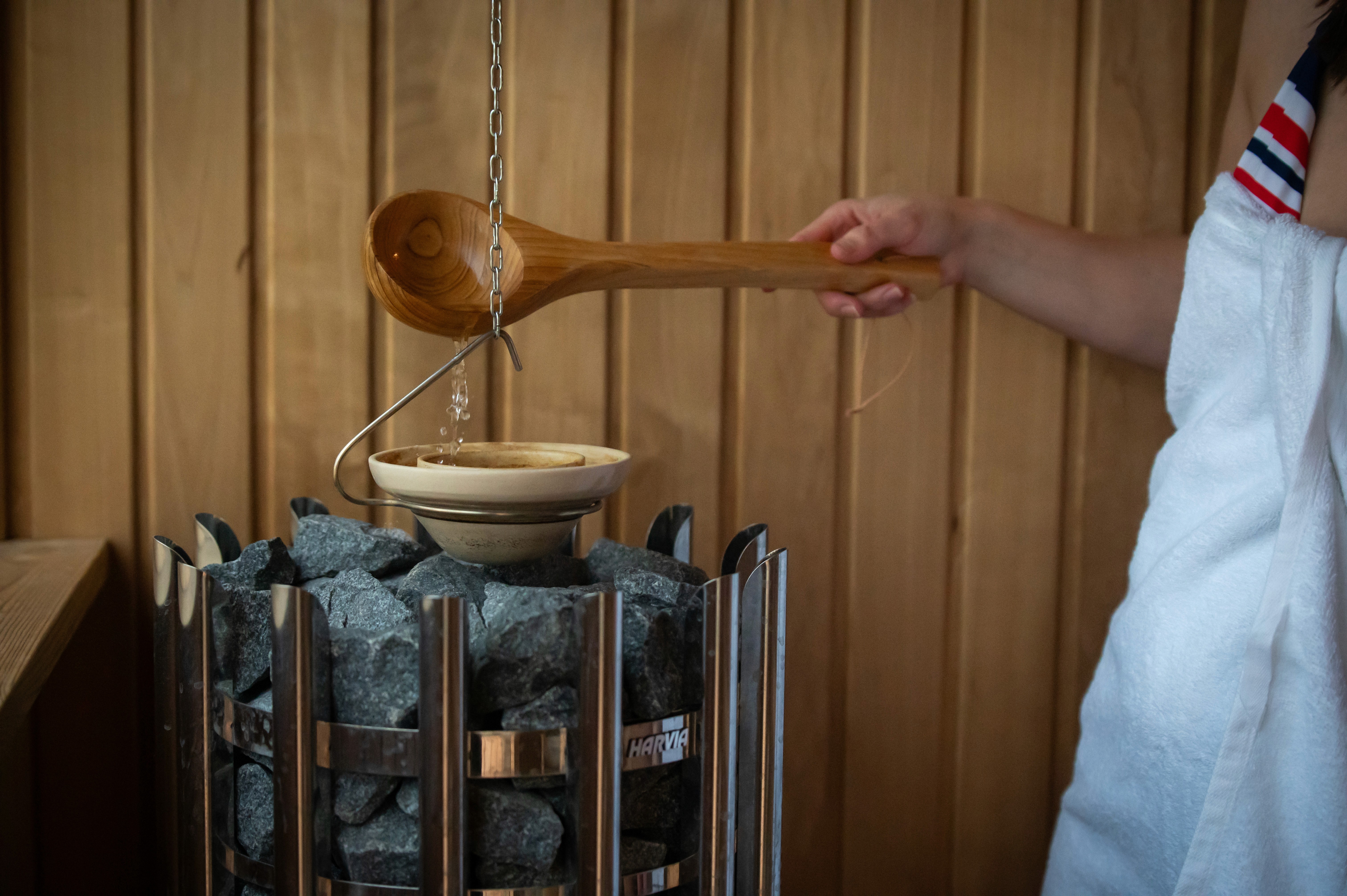 Egy fehér törölközőt viselő személy fa merőkanállal vizet önt a forró szaunakövekre, miközben a háttérben fa burkolat látható, és gőz keletkezik.