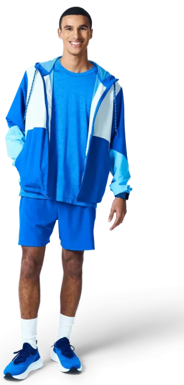 A young man stands confidently in a blue athletic outfit, including shorts and a hooded jacket.