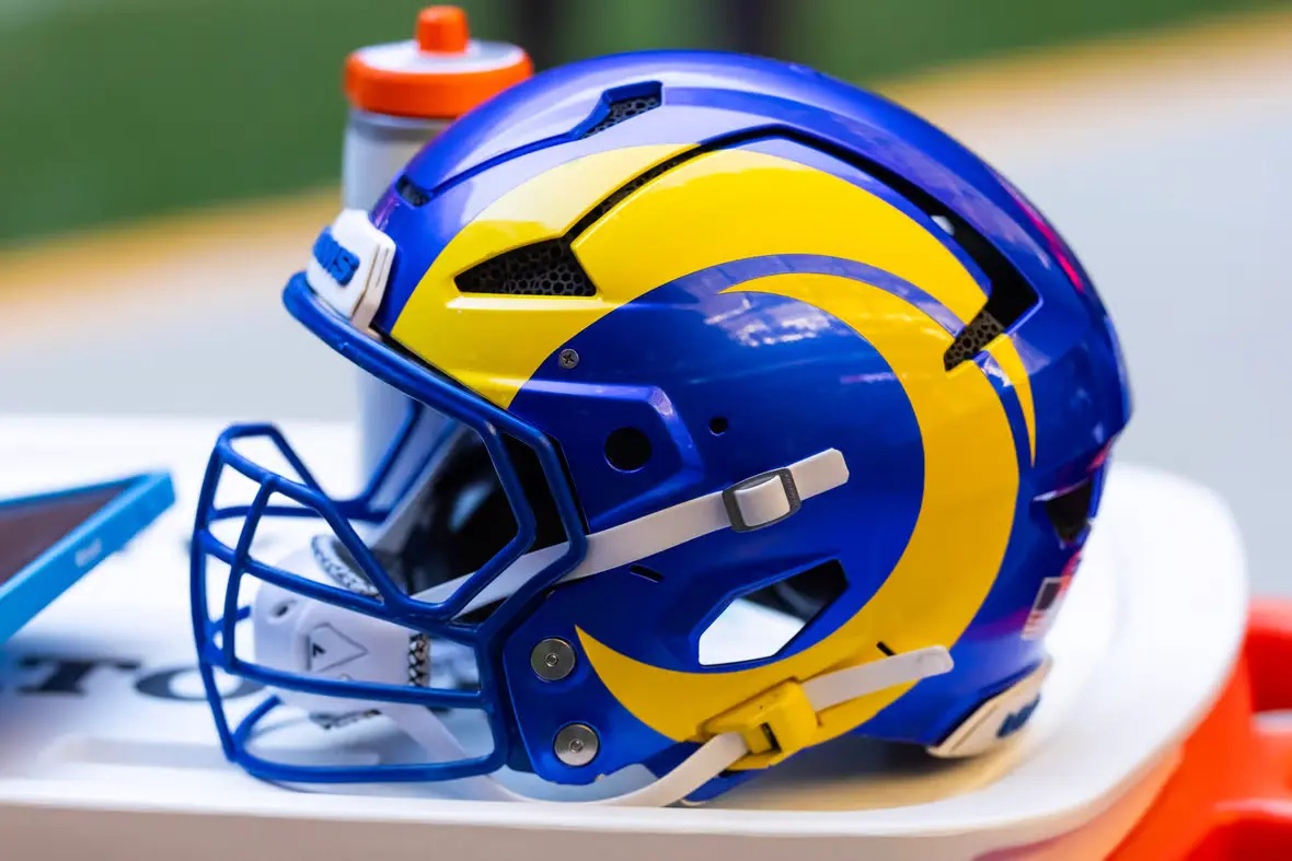 Close up of a Los Angeles Rams football helmet in blue and yellow resting on a bench with equipment in the background.