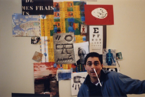 A photo of a person in front of a wall filled with different artwork like picture of hieroglyphs, maps, eye control chart letters, hand imprints and others.