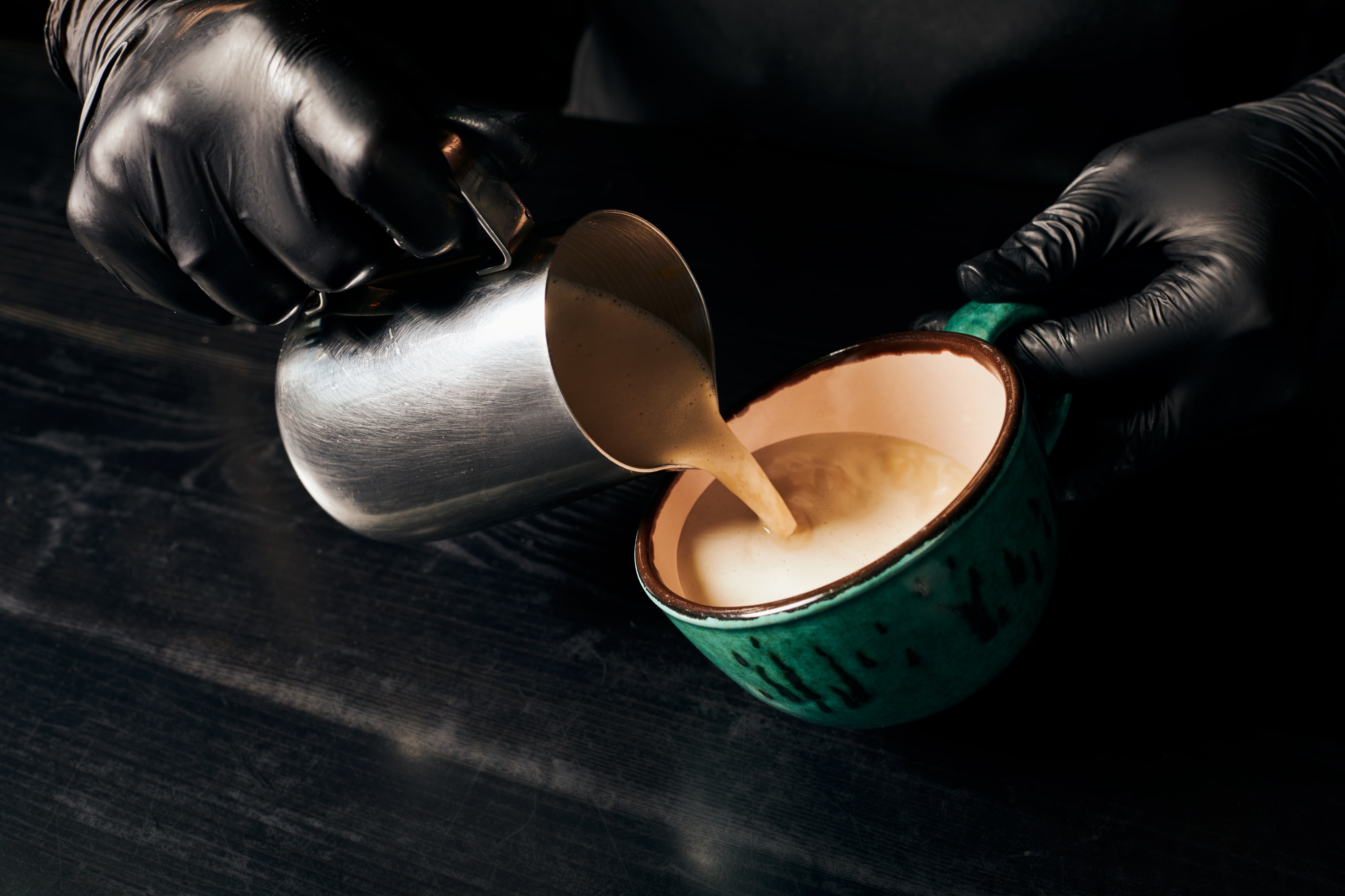Barista pouring steamed milk from a metal pitcher into a ceramic cup, in a dark, moody café close-up.