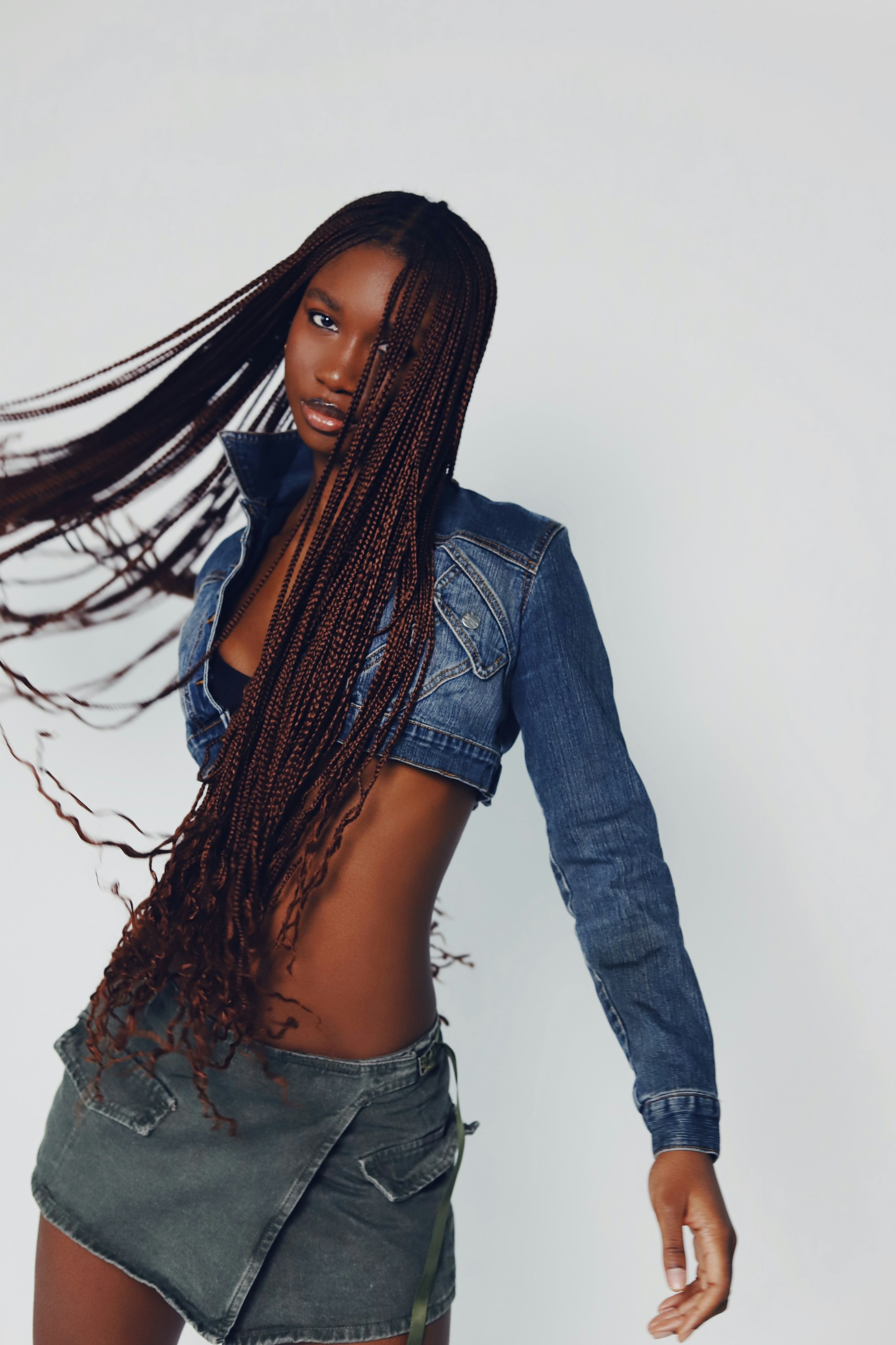 A dynamic shot of the woman from the waist up as she moves, sending her long brown braids flying. She is wearing a cropped denim jacket and a green utility-style skirt against a plain white background.