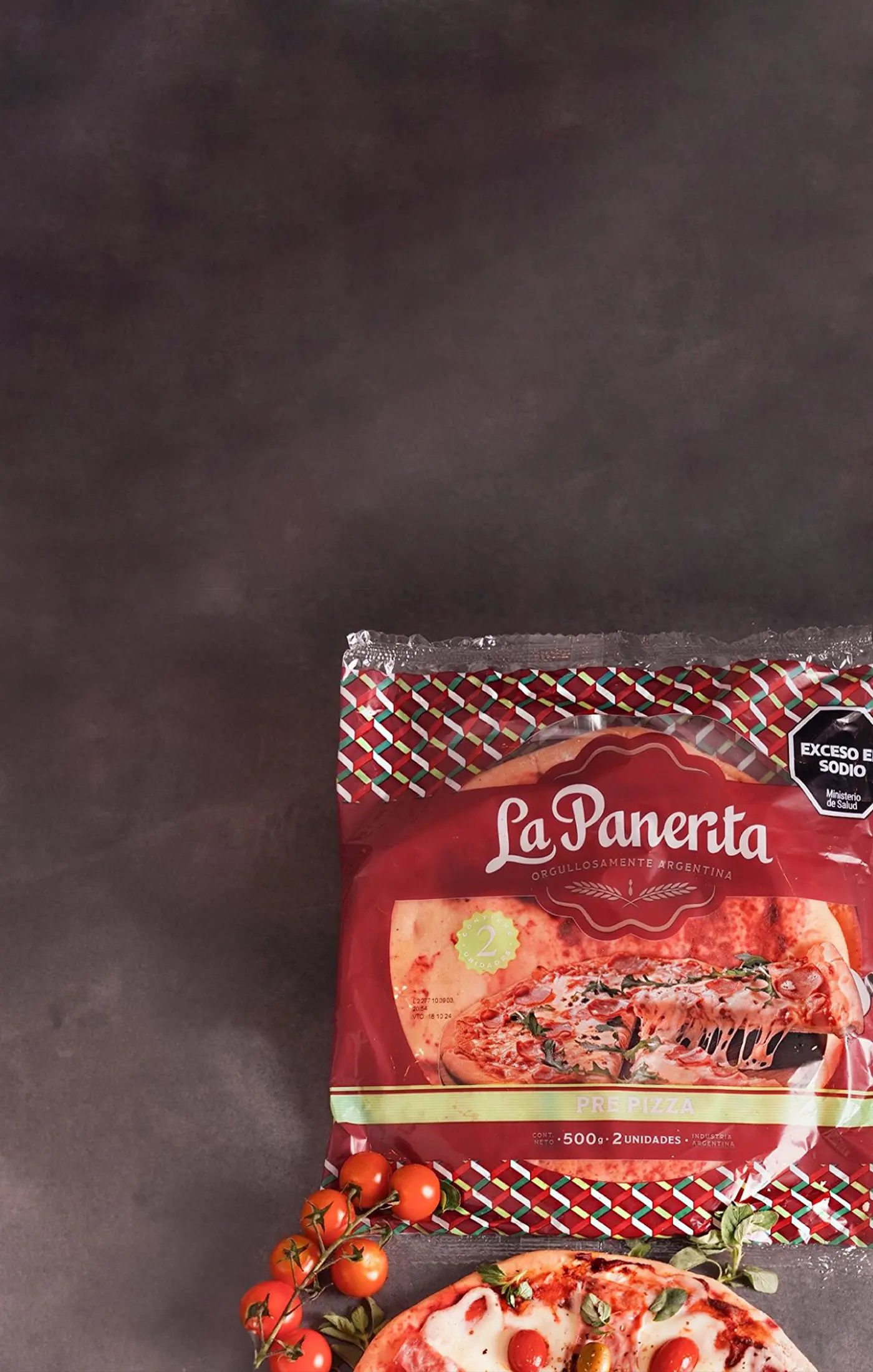 Packaged pizza labeled "La Panerta" on a dark surface, accompanied by fresh vine tomatoes and a partially visible pizza. The mood is appetizing and rustic.