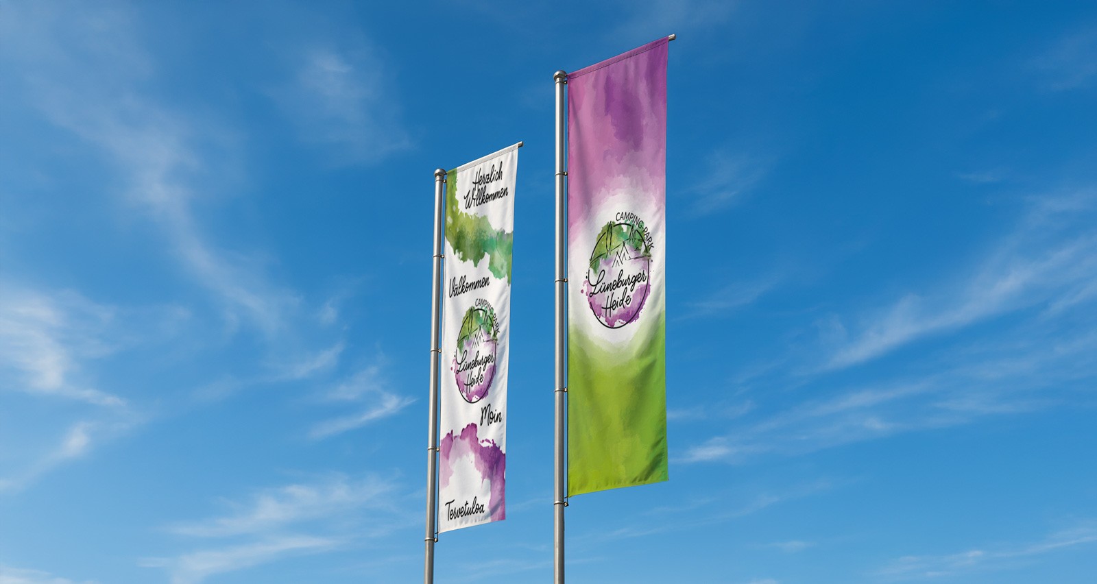 Zwei bunte Fahnen mit Aquarellhintergrund und einem kreisförmigen Logo flattern im Wind vor einem strahlend blauen Himmel mit Wolkenfetzen. Auf der linken Flagge stehen die Worte Herzlich Willkommen in 4 verschieden Sprachen. Auf der rechten Flagge ist nur das Aquarell mit dem Logo des Campingparks zu sehen.