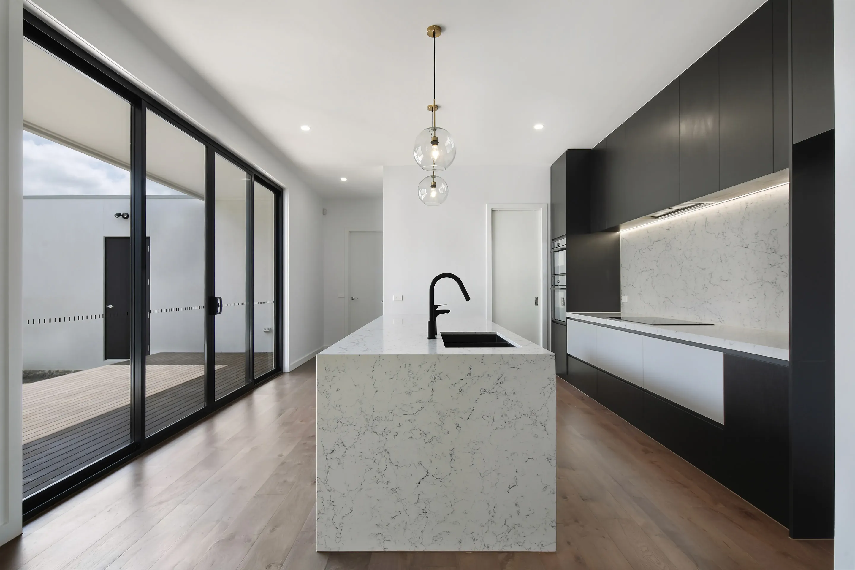 Kitchen and living with a veined white stone waterfall island, glass globe pendants, black overhead cabinetry with white base units, black tap and black-framed sliding doors to a courtyard