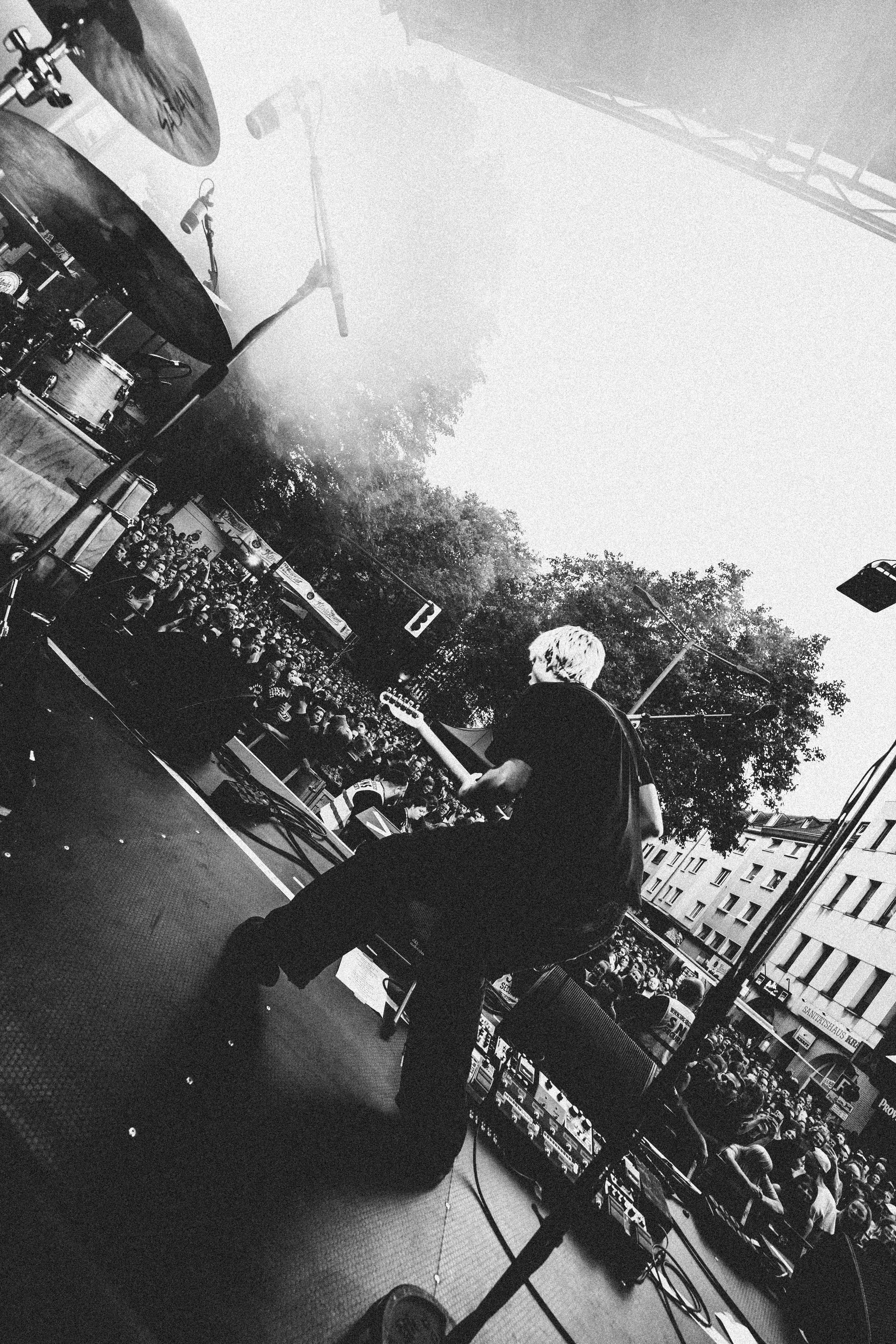 Schwarz-weiß-Aufnahme eines Gitarristen auf einer Open-Air-Bühne, der mit dem Rücken zur Kamera spielt und auf ein großes, dicht gedrängtes Publikum blickt. Links im Bild sind Teile des Schlagzeugs zu sehen, im Hintergrund Bäume und Hausfassaden.