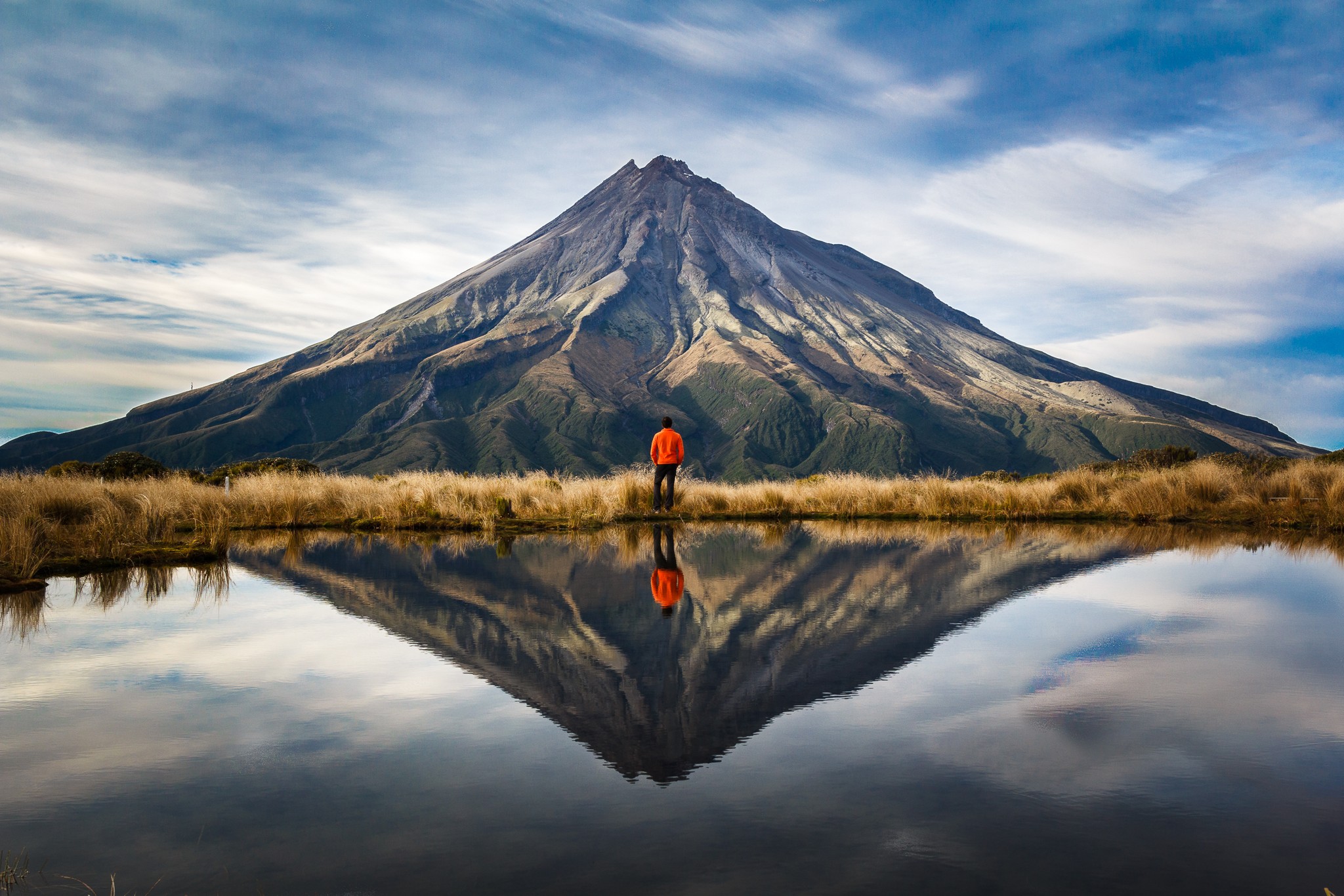 A man standing at the edge of a lake looking toward a mountain that's reflected on the water