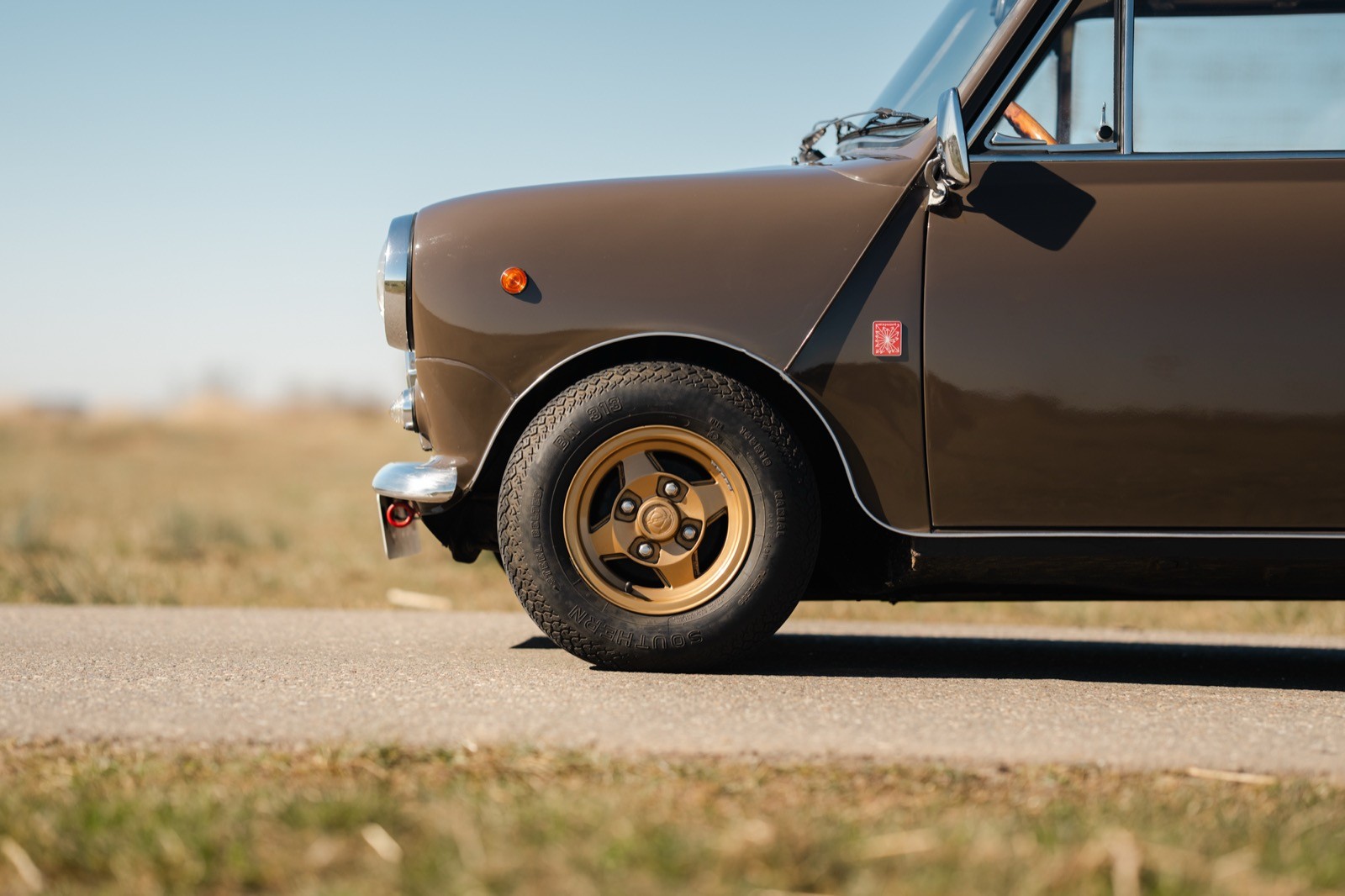 A close-up view of a vintage car's side, showcasing a brown body and a golden wheel against an open field backdrop.
