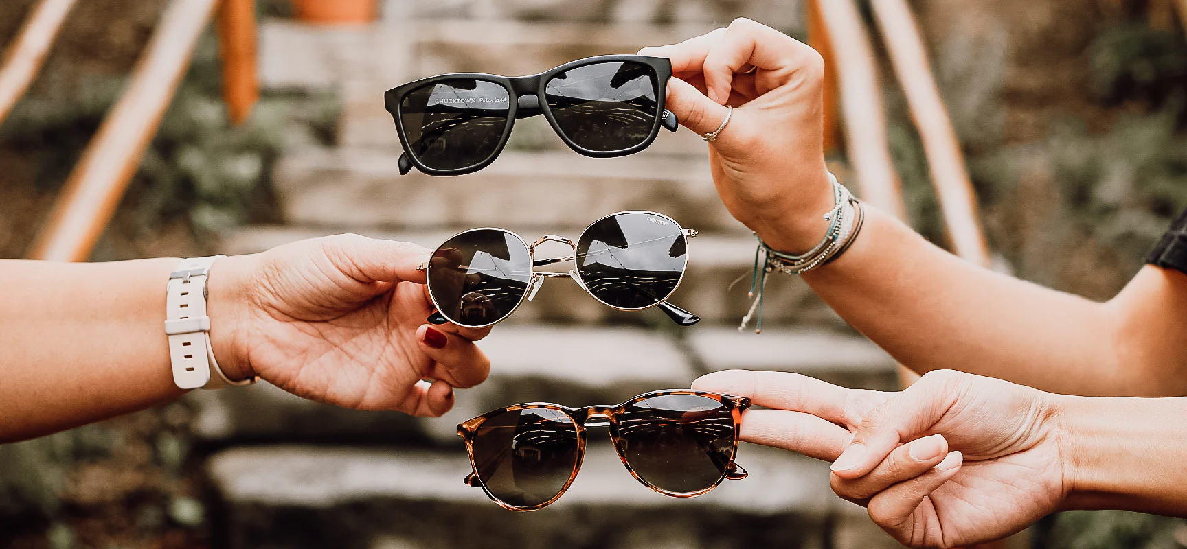 Hands holding various pairs of sunglasses against a rustic background, showcasing different styles.