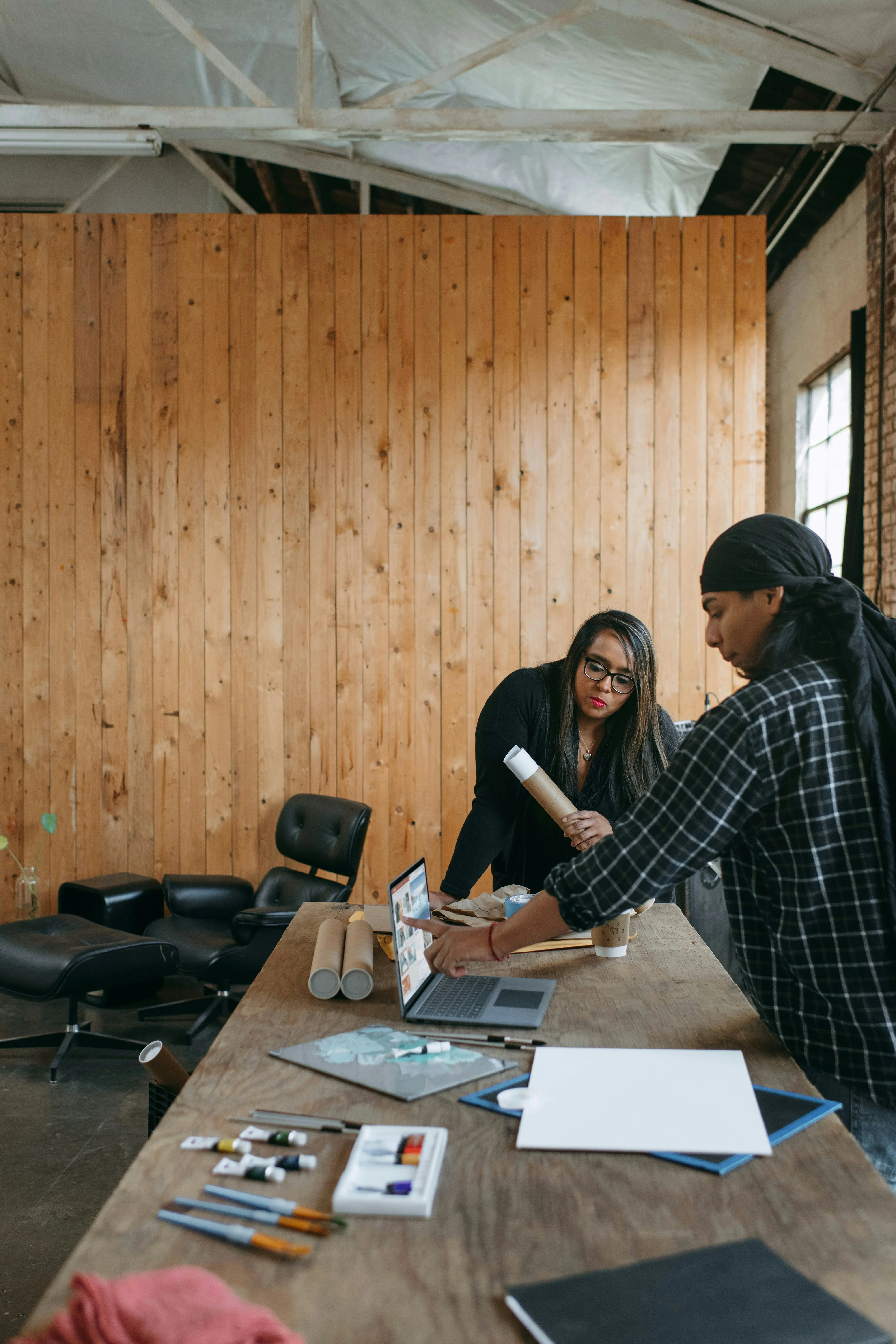 Two people working at a table in an informal setting with wooden walls, laptops, and papers around.