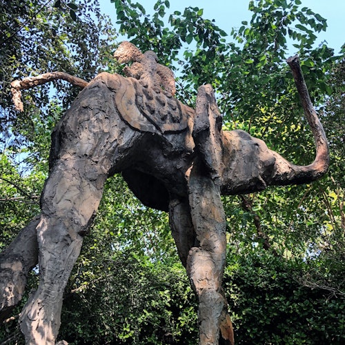 Isang iskultura ng elepante na gawa sa putik na may nakasakay na tao, napapalibutan ng malalago at berdeng mga halaman at puno.