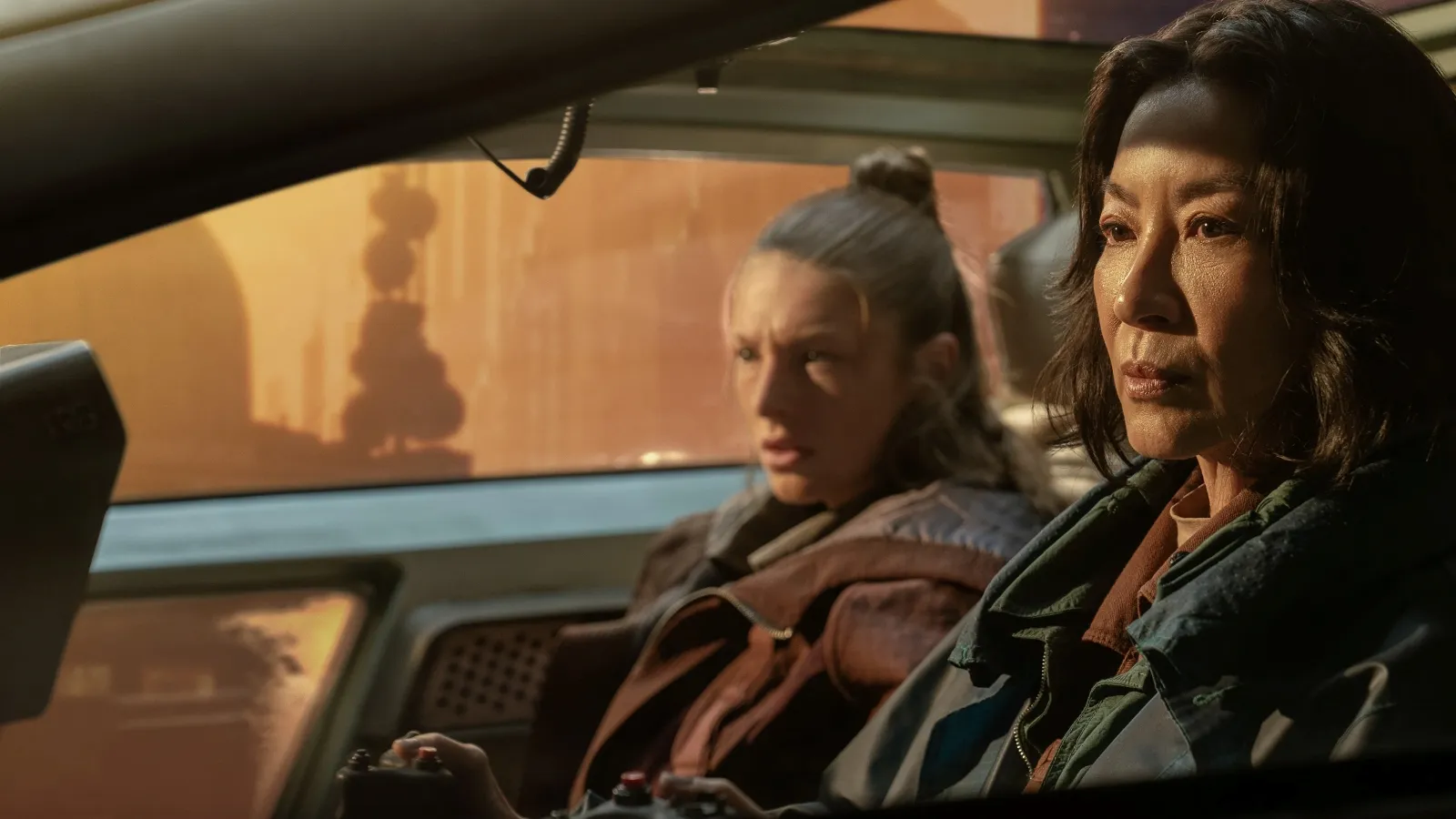 Two individuals sit in a futuristic vehicle with a high-tech dashboard, gazing intently at an orange-tinted futuristic cityscape through the windshield as sunlight casts dramatic shadows inside the cabin.