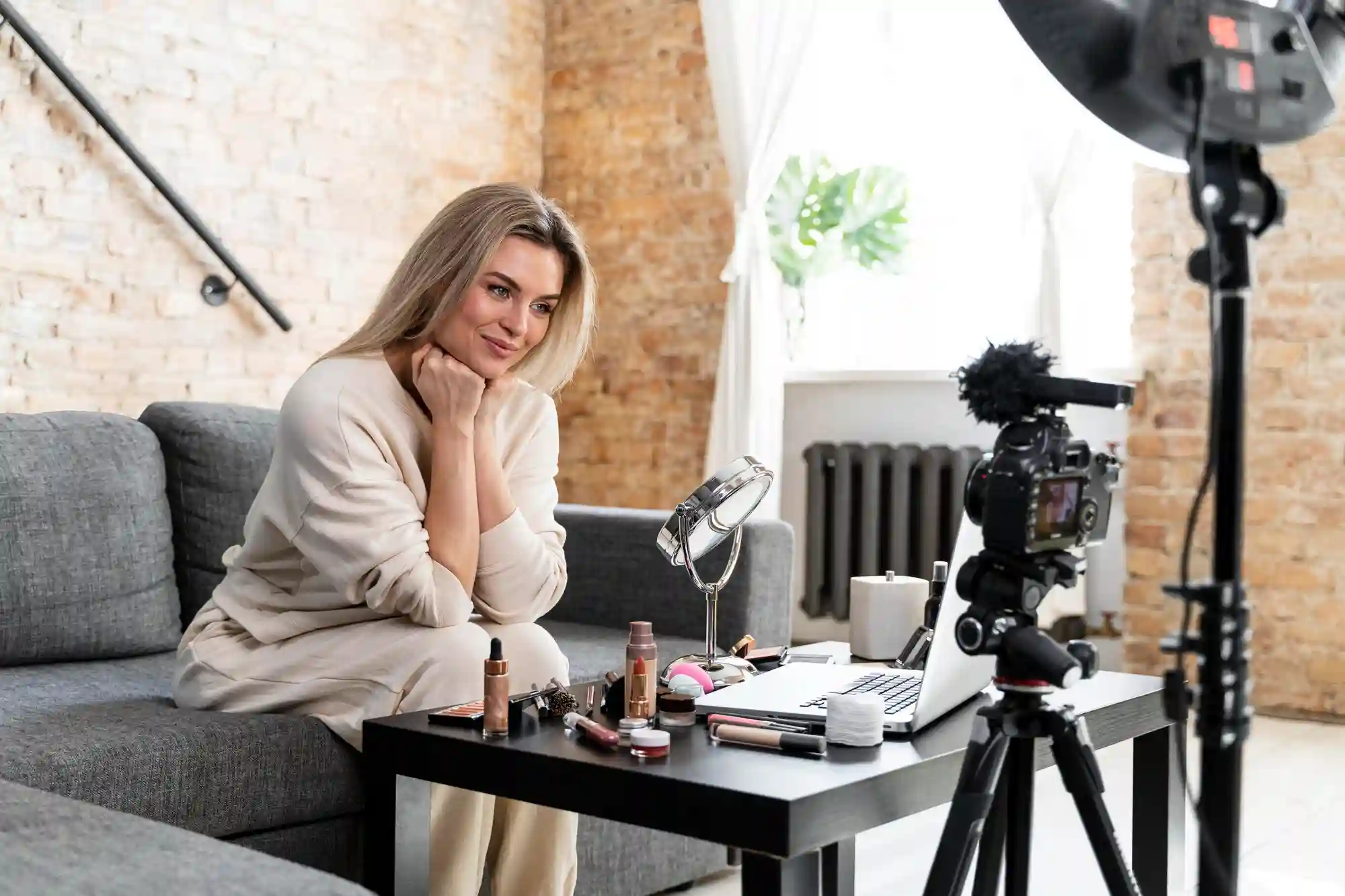 Blonde woman posing on a couch for a makeup tutorial video setup with a camera and lighting equipment.