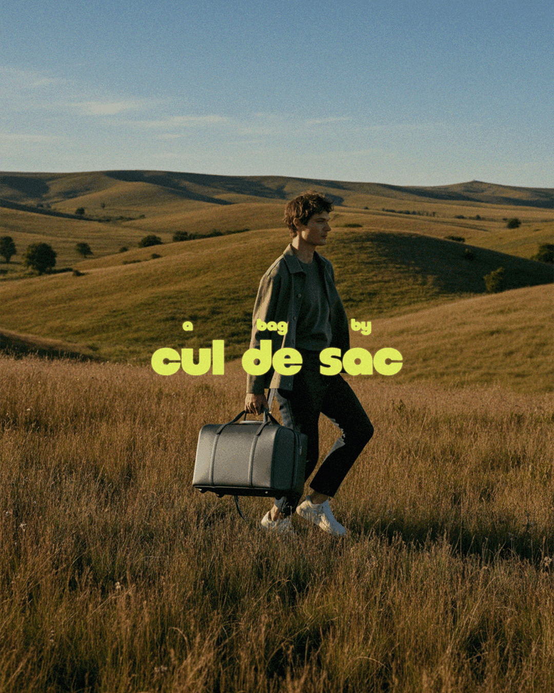 A person walking through open grassland carrying a large dark duffel bag, with the cul de sac wordmark overlaid in chartreuse