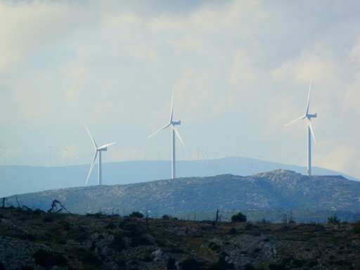 Wind turbines stand on a distant hill under a cloudy sky, surrounded by a rugged landscape.
