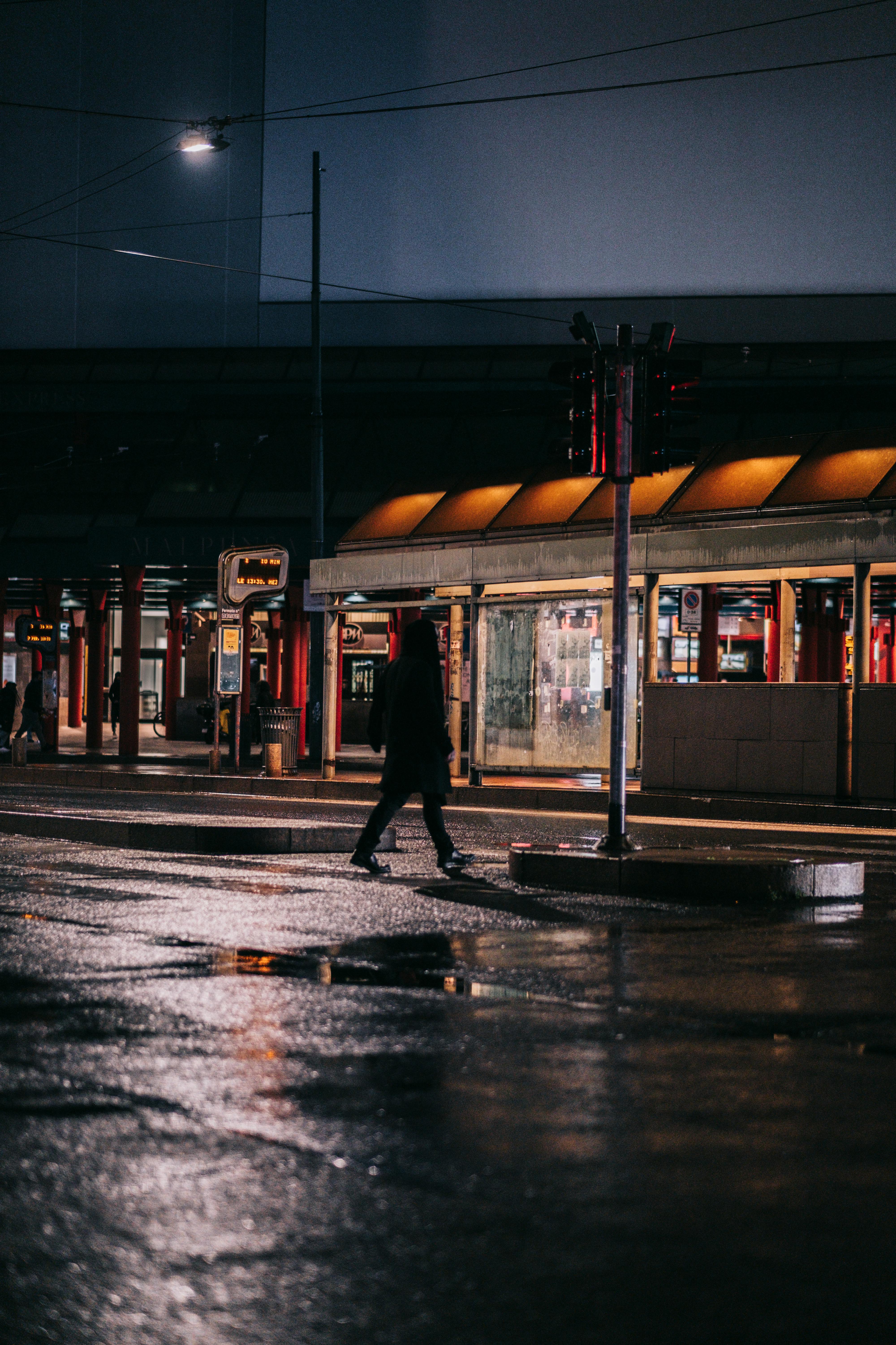 Urban night photography of a silhouetted pedestrian walking past a brightly lit bus shelter with a warm orange awning, reflecting on the wet city street.
