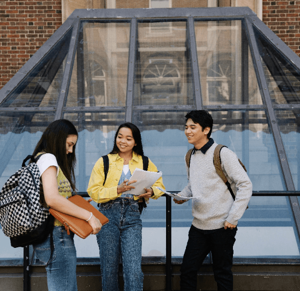 Three smiling students with backpacks and folders stand outside a brick building in front of a glass entrance.