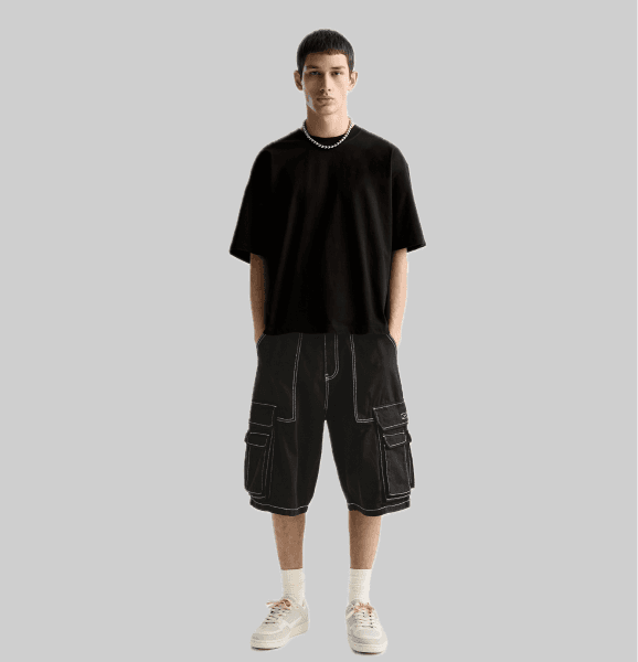 "KUGO mobile fashion app displaying a male model wearing a black oversized t-shirt with a chain necklace and black cargo pants with contrast stitching. Modern streetwear style on a sleek smartphone screen."
