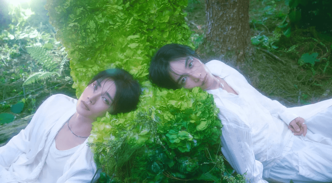 JEONGHAN and JOSHUA in white, resting against green foliage.