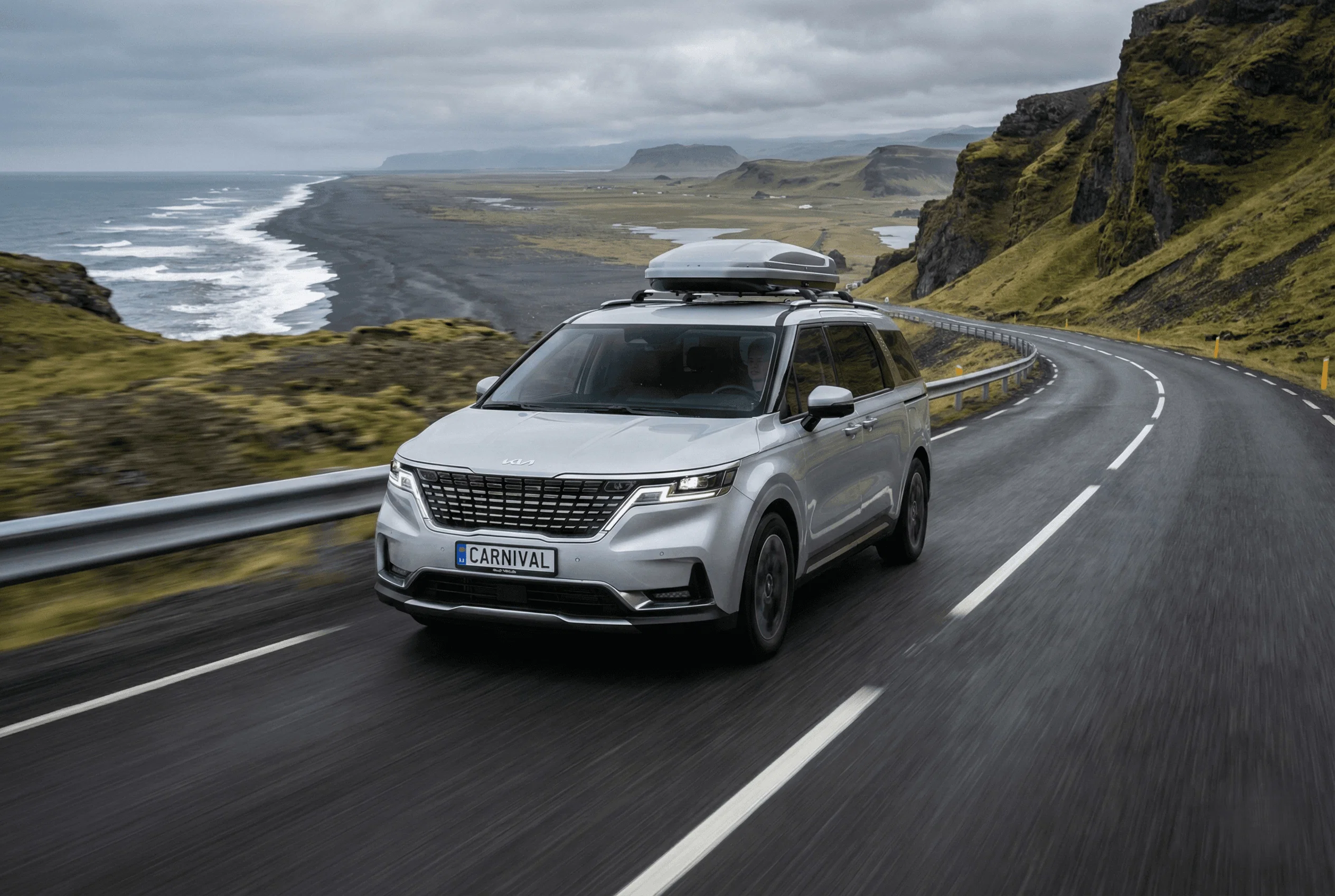 A silver Kia SUV with a roof box driving along a winding coastal road near black cliffs.
