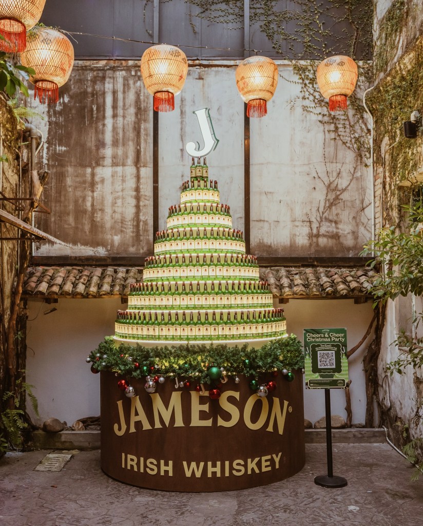 A festive display featuring a pyramid made of Jameson whiskey bottles, adorned with Christmas decorations and lights, with a backdrop of traditional architecture and red lanterns.